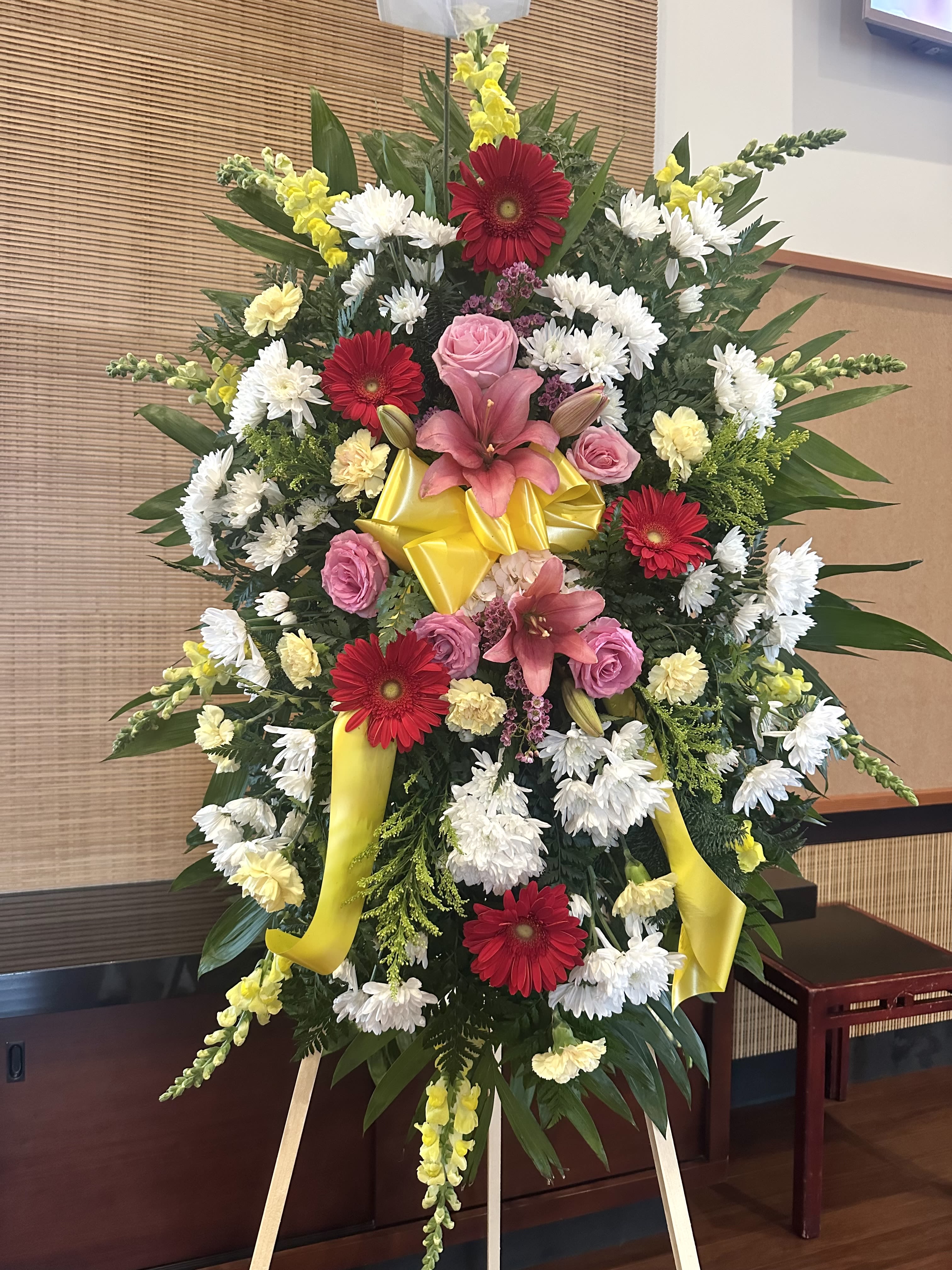 Standing floral wreath with pink lilies, red gerberas, white daisies, and yellow ribbon