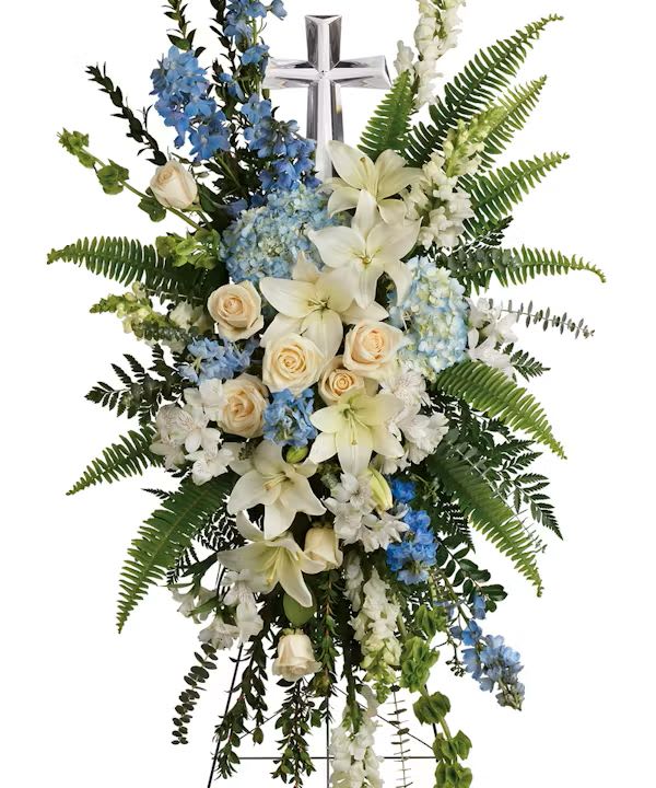 Standing funeral spray with white lilies, roses, and blue flowers on a cross easel