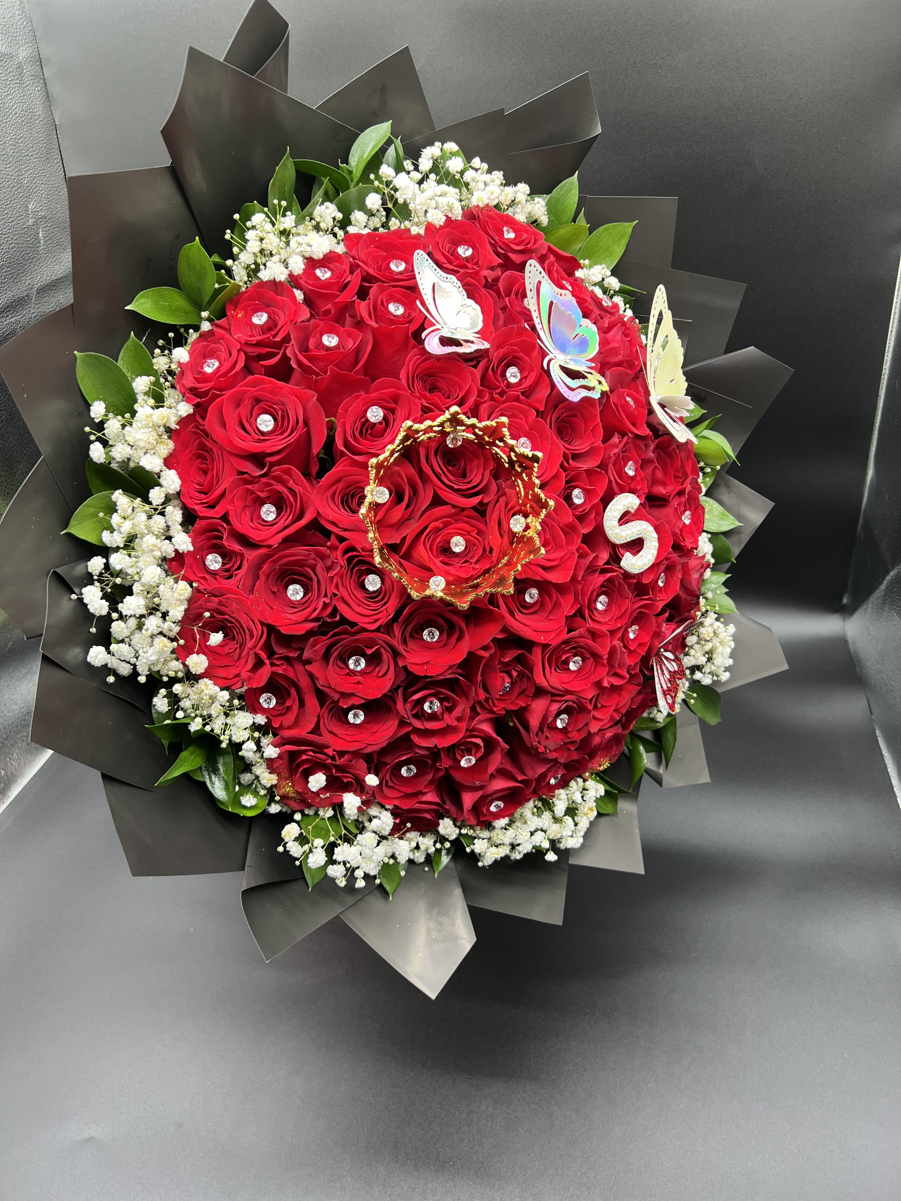 Tightly packed bouquet of red roses with jeweled centers wrapped in black paper with white filler flowers