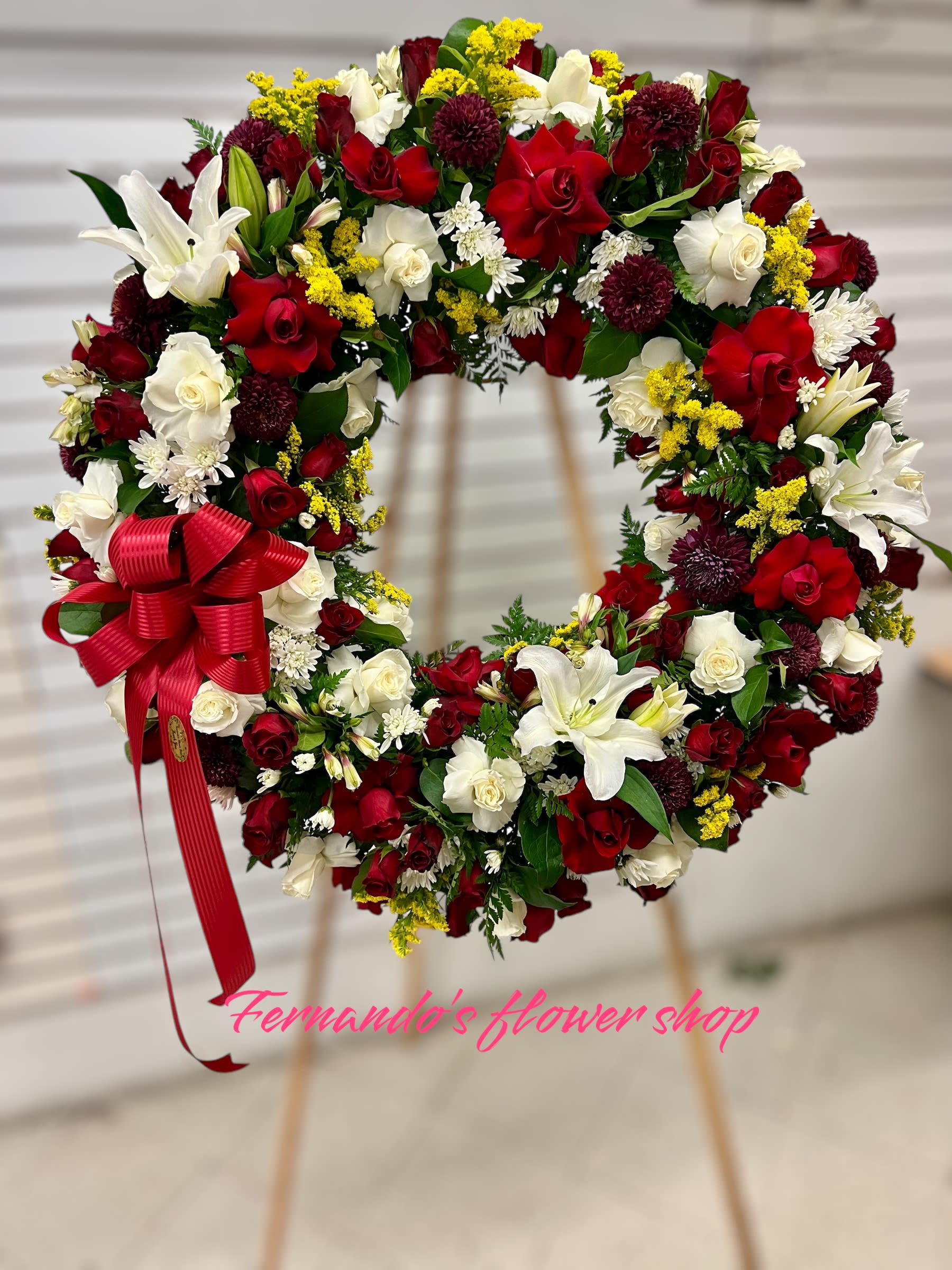 Standing wreath of red and white roses with white lilies, mums, and a red bow on an easel.