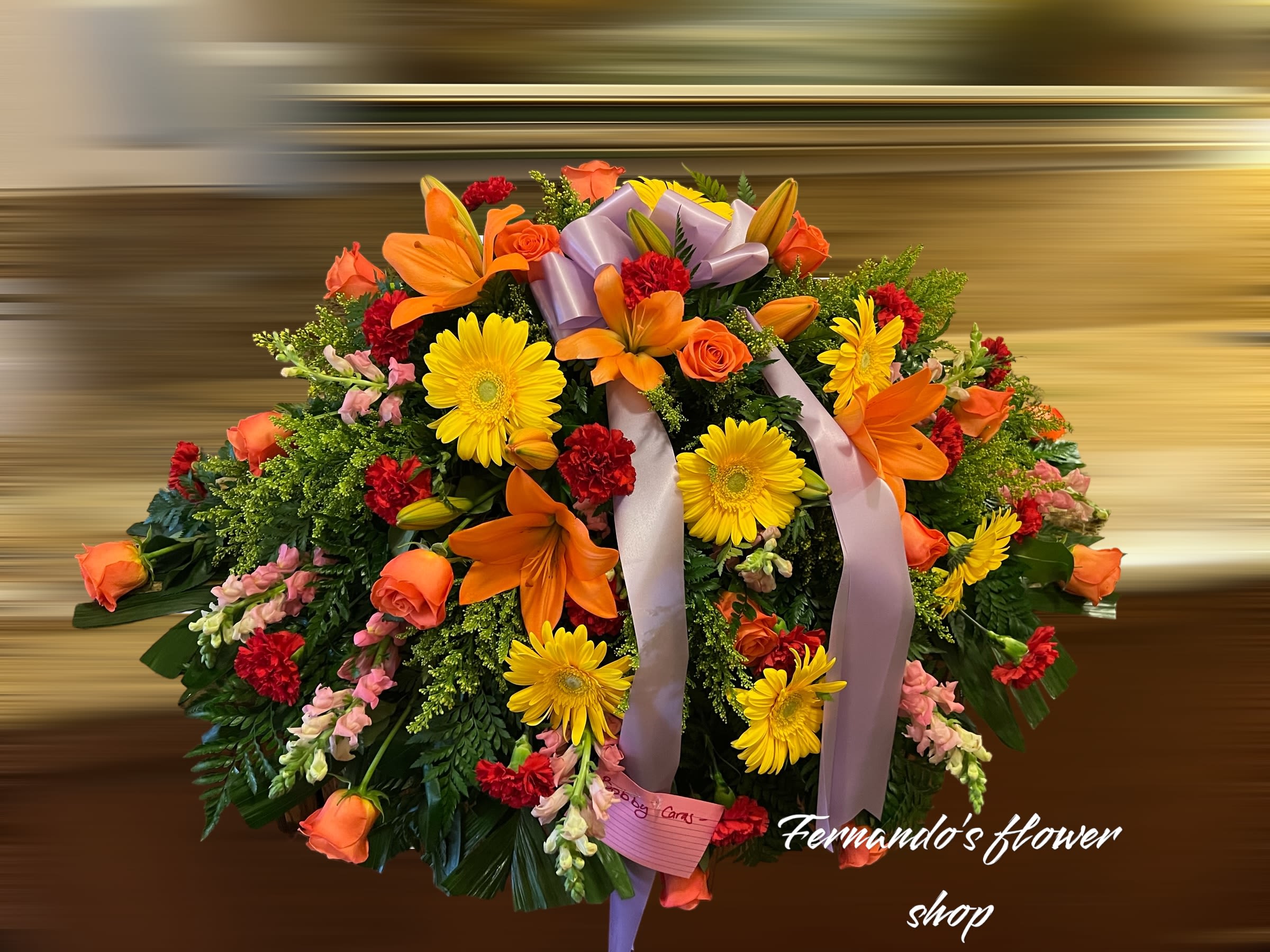 Large round spray of orange lilies, yellow gerberas, roses, and red carnations with a lavender bow.