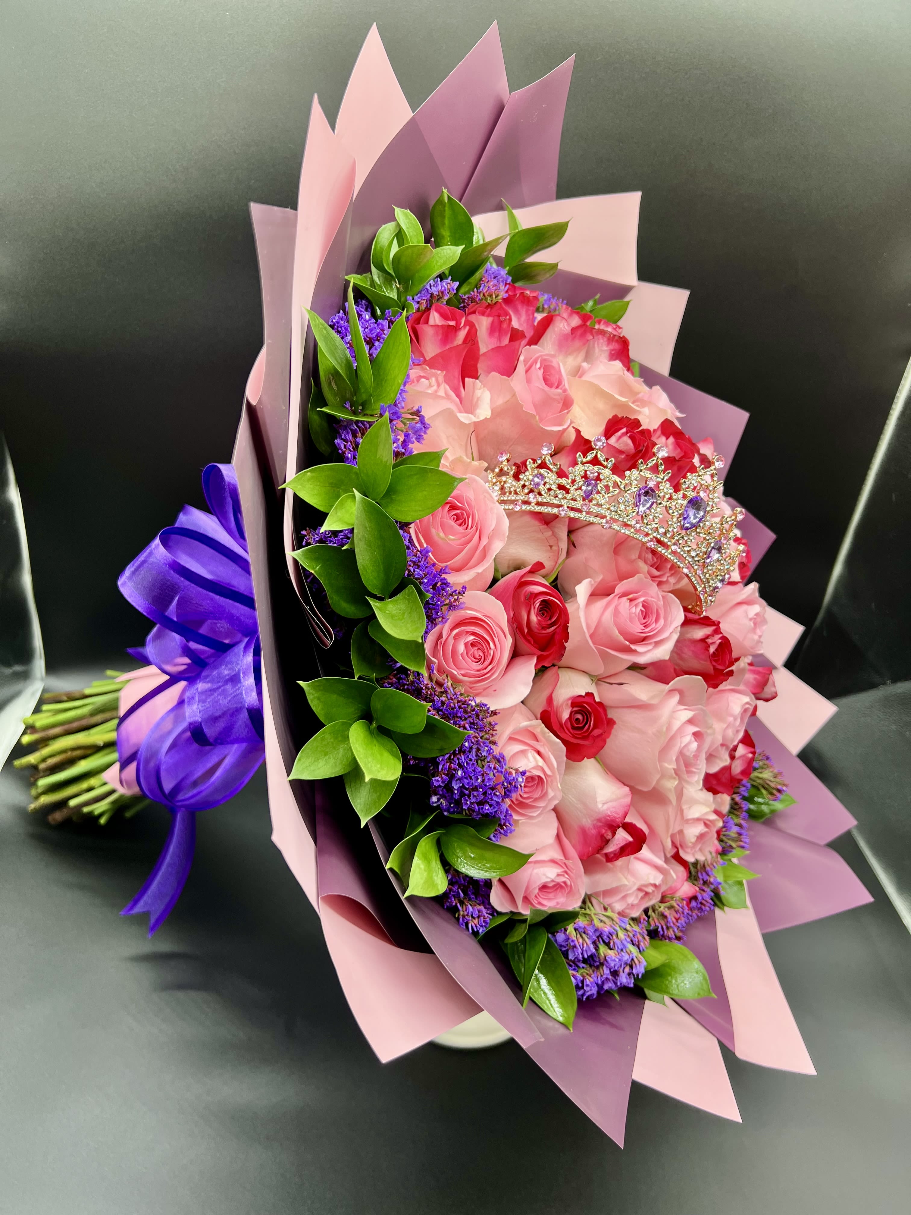 Bouquet of pink and red roses with purple filler flowers, wrapped in pink paper with a small tiara decoration.