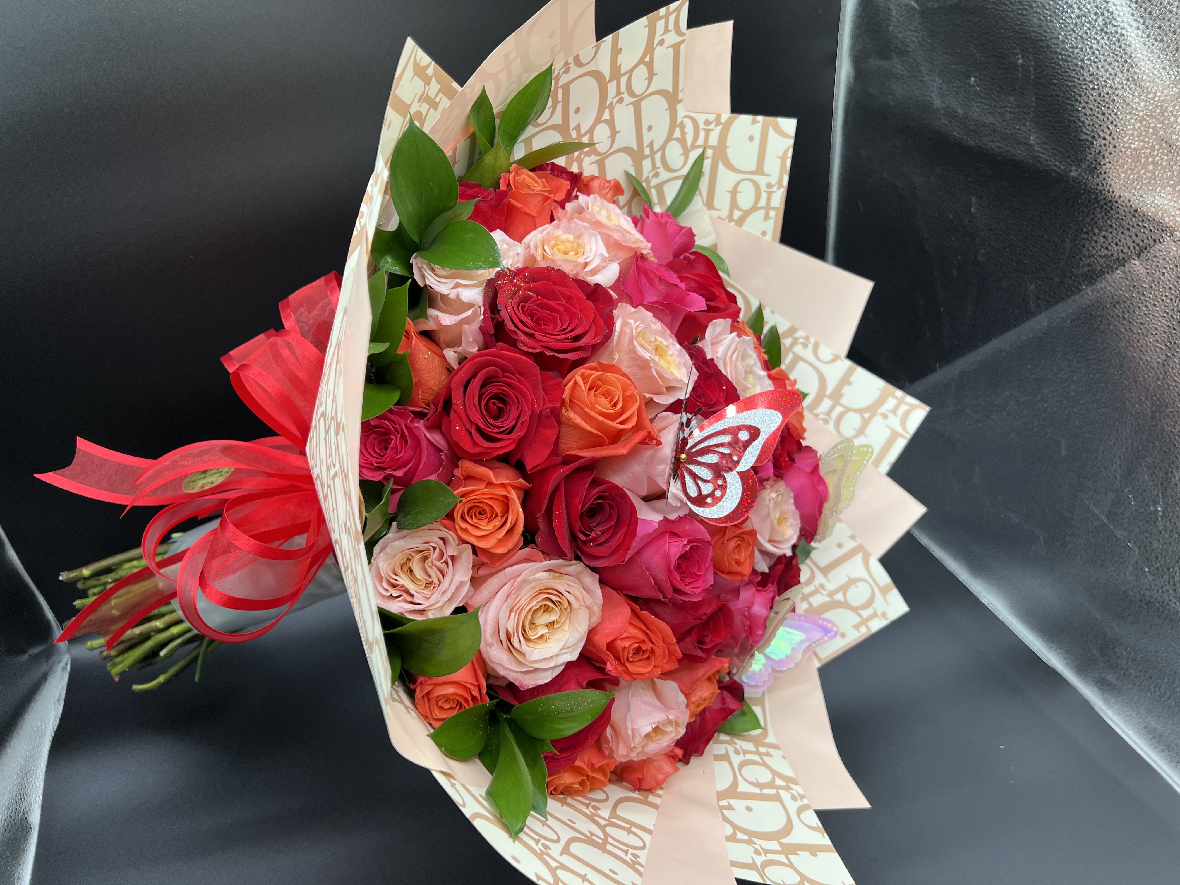 Hand-tied bouquet of red, pink, and peach roses wrapped in patterned paper with a red bow and butterfly accents