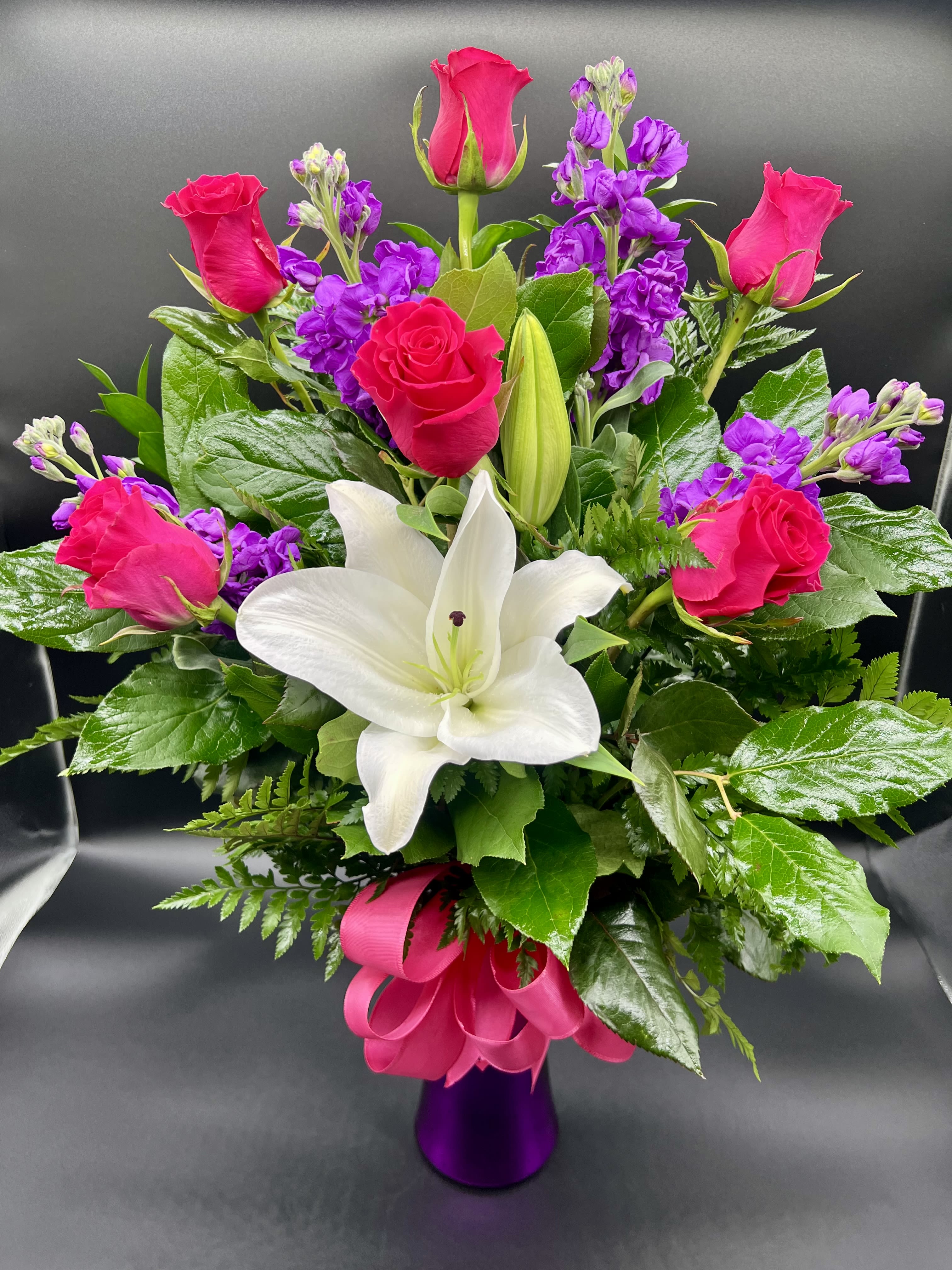 Arrangement of hot pink roses, purple stock, and white lilies in a purple vase with a pink bow
