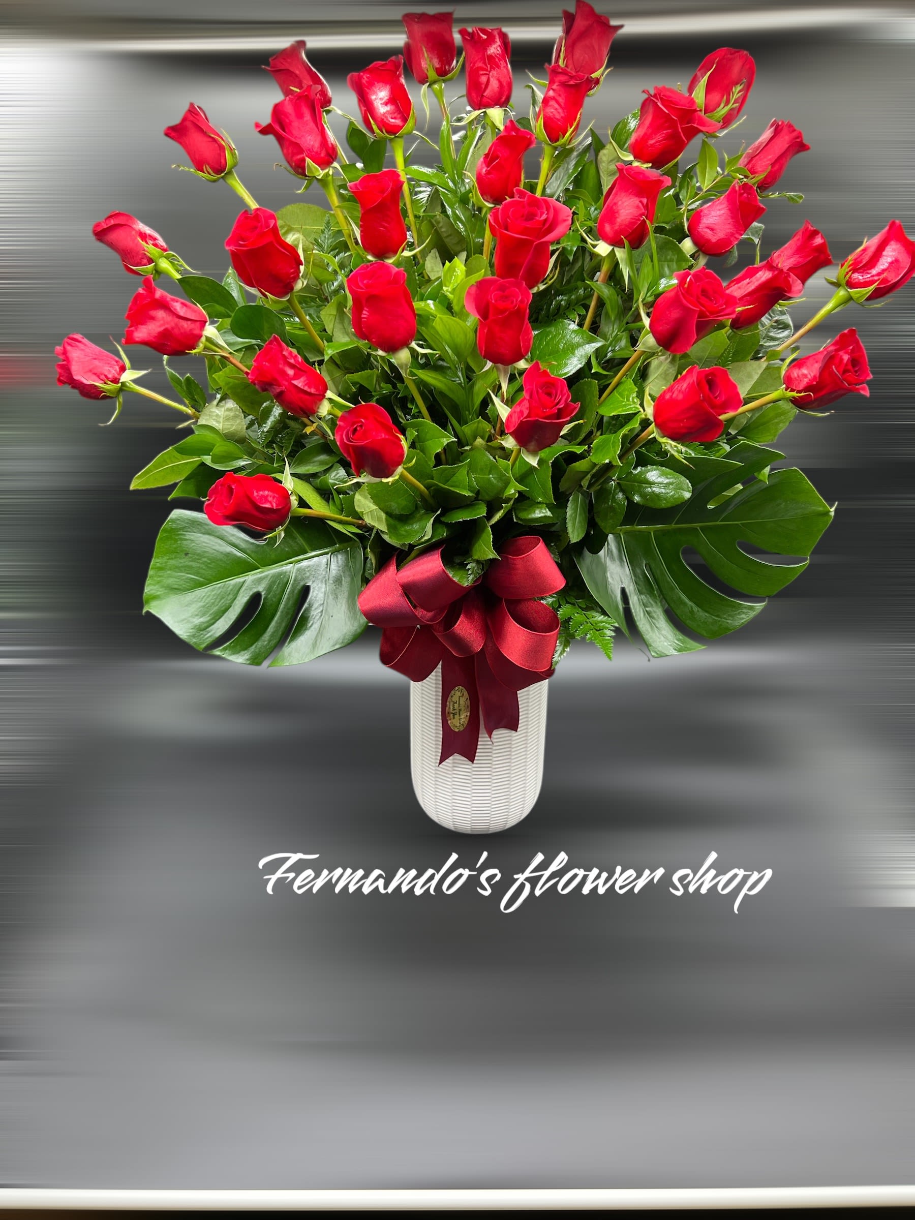 Tall arrangement of red roses in a white vase with large glossy leaves and a red ribbon bow