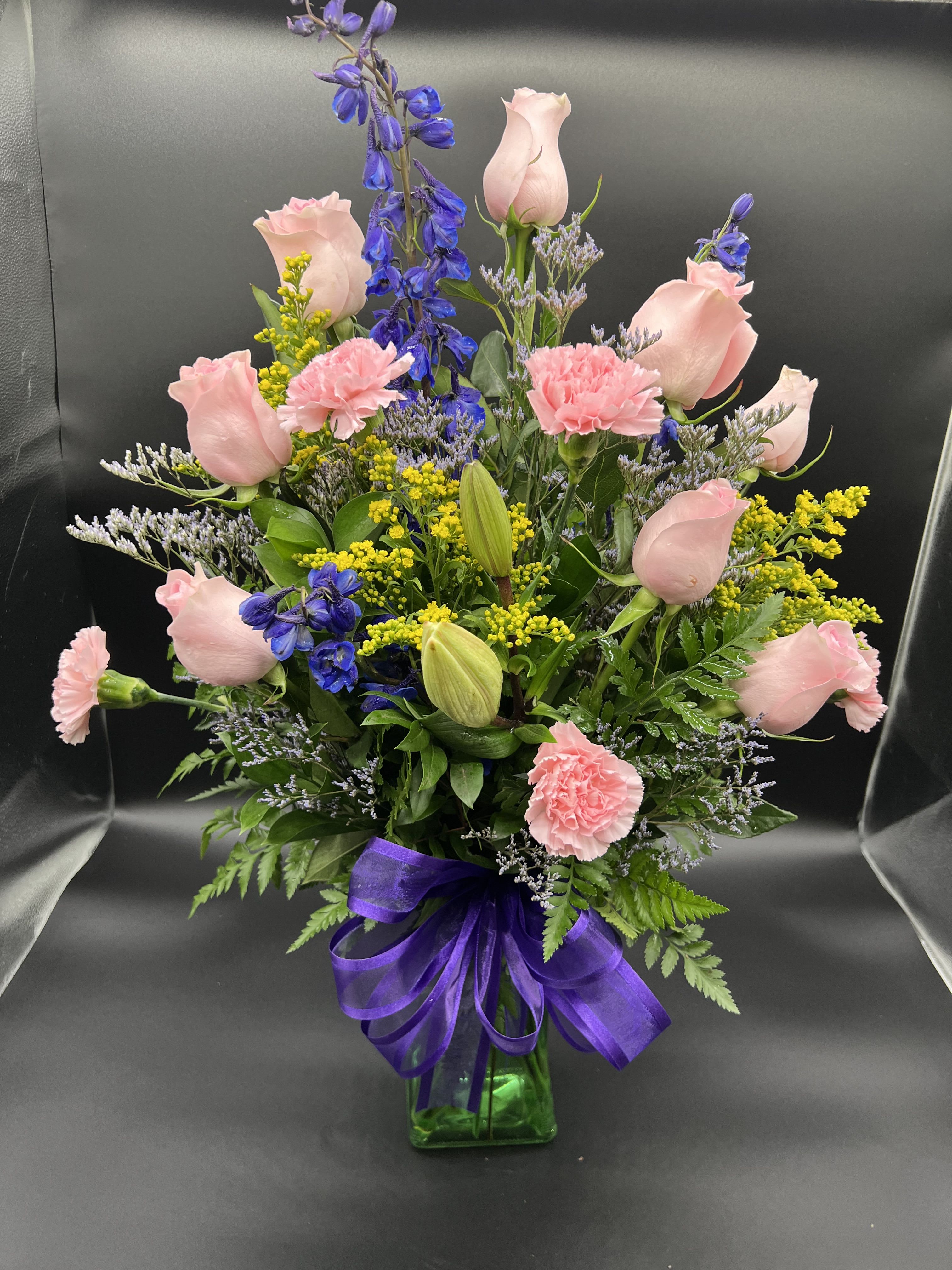 Tall mixed bouquet of pink roses and carnations with blue delphinium in a green vase with purple bow