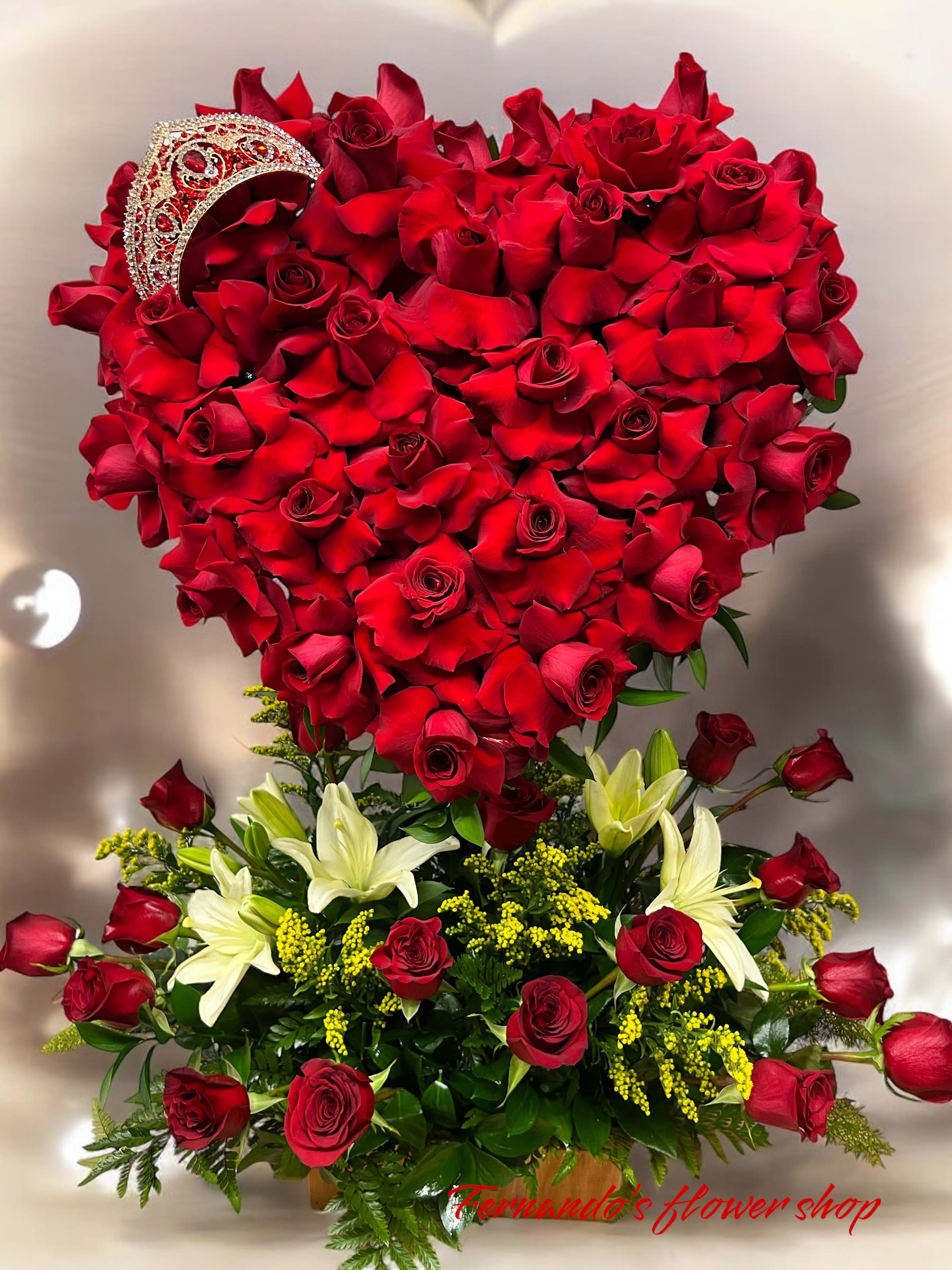 Heart-shaped red rose display with tiara above a base of red roses, white lilies, and yellow filler flowers.