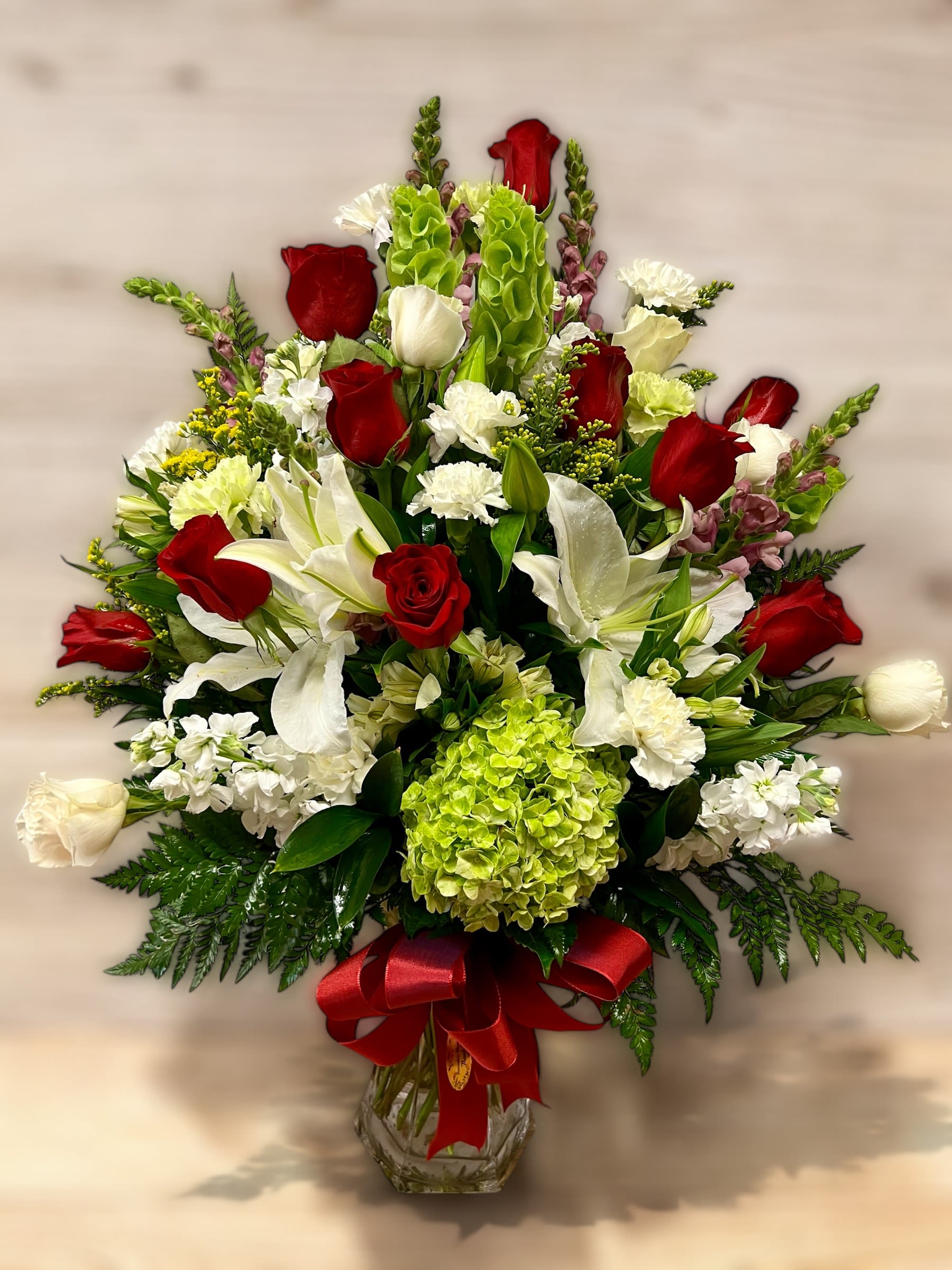 Tall red and white arrangement with roses, lilies, and green hydrangea in a glass vase with red bow.