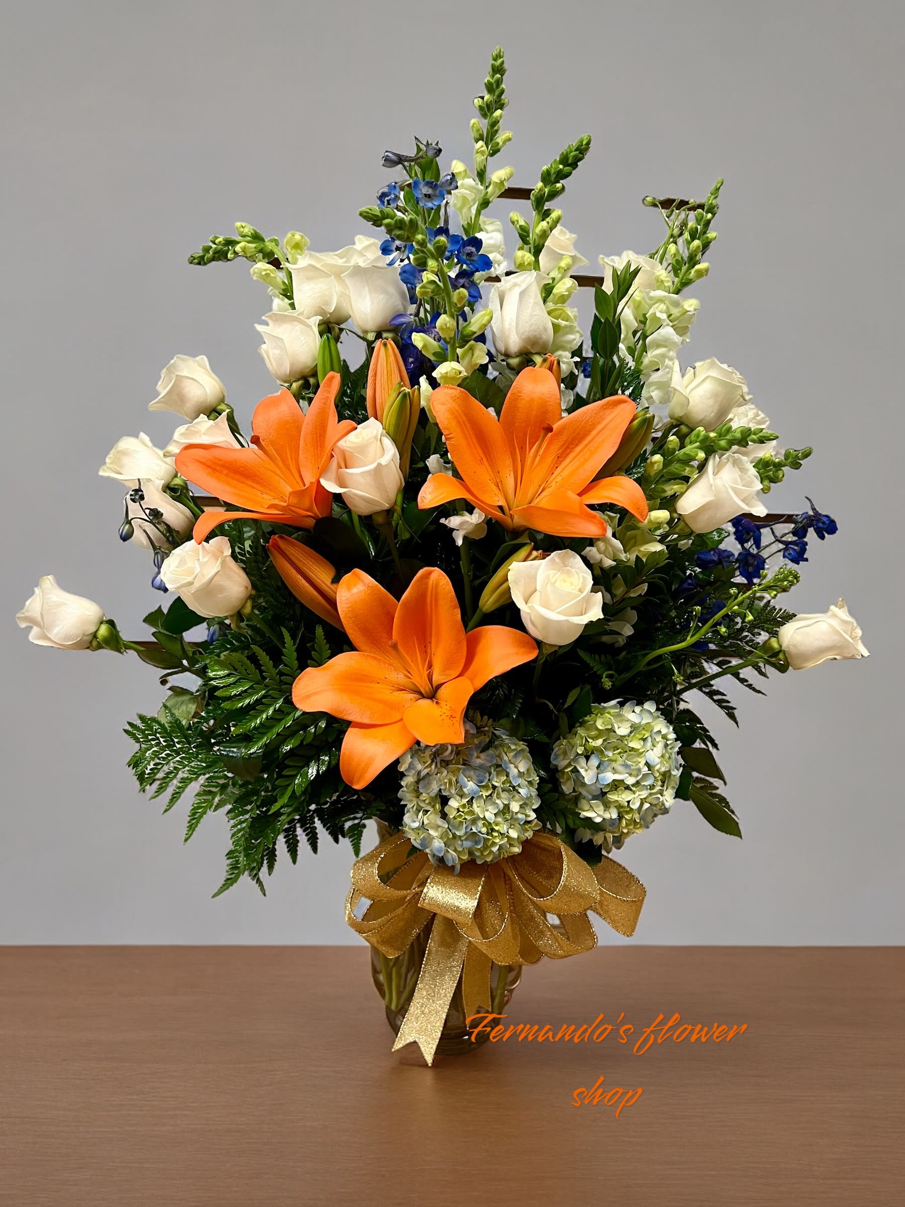 Tall arrangement of orange lilies, white roses, blue flowers, and hydrangeas in a gold vase with a bow