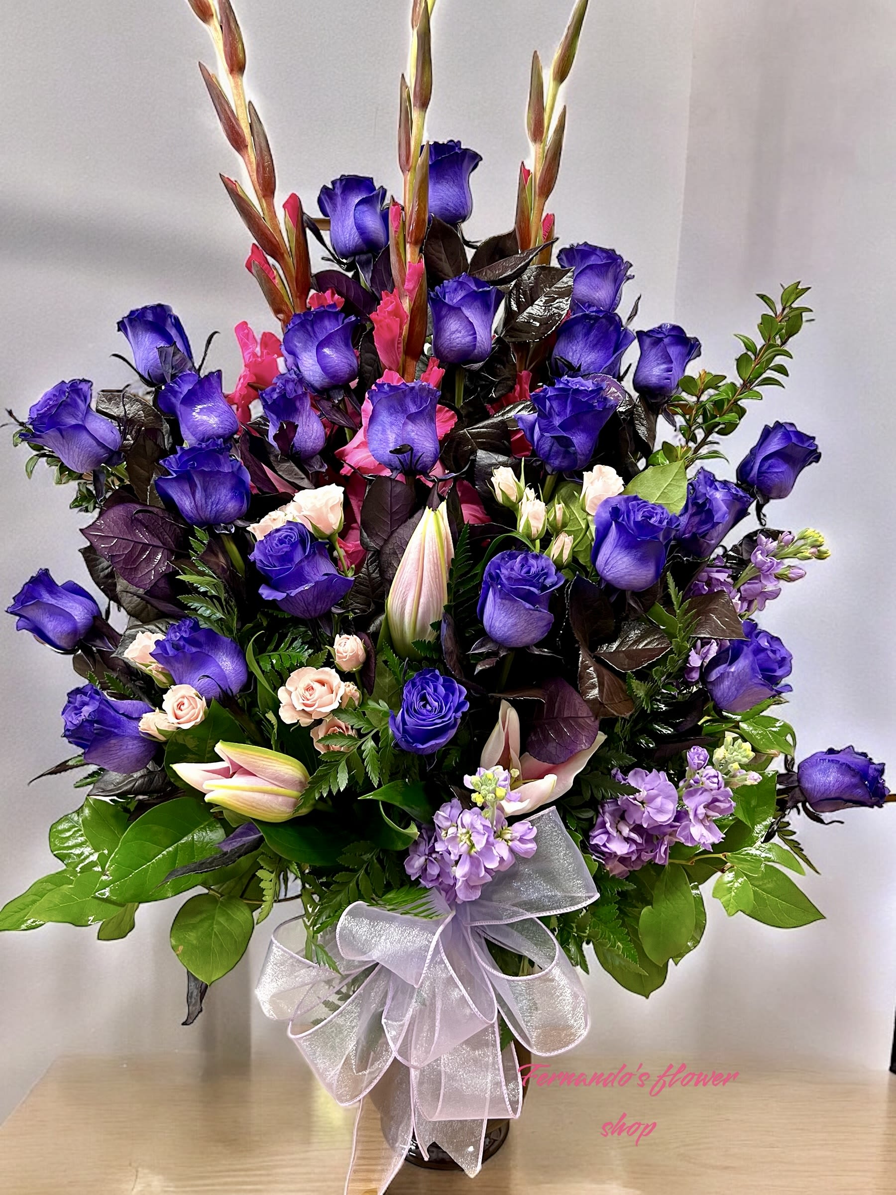 Tall arrangement of purple roses, pink lilies, and mixed blooms in a vase with a sheer ribbon bow