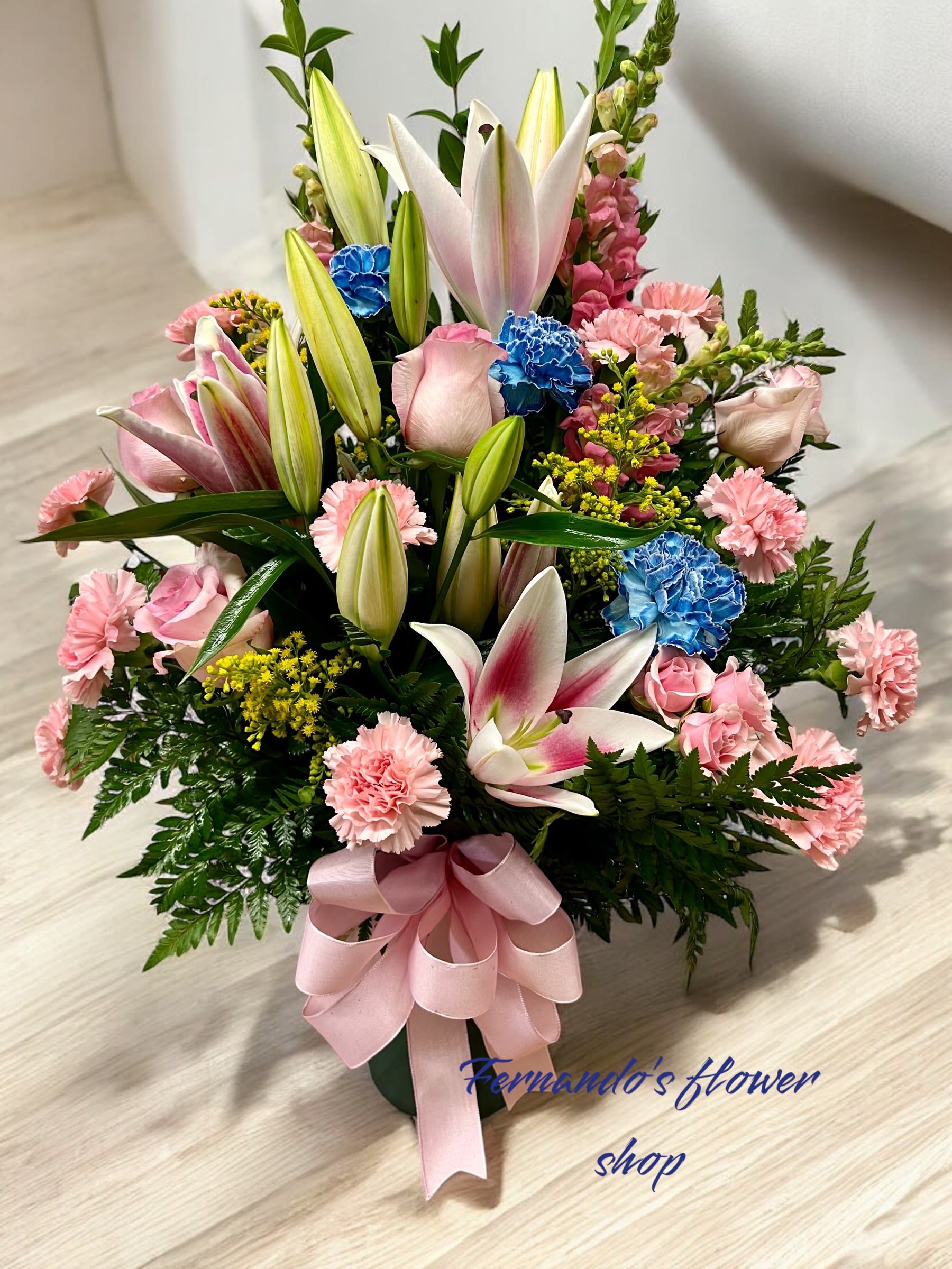 Tall arrangement of pink lilies, roses, carnations, and blue flowers with a pink bow in a green vase