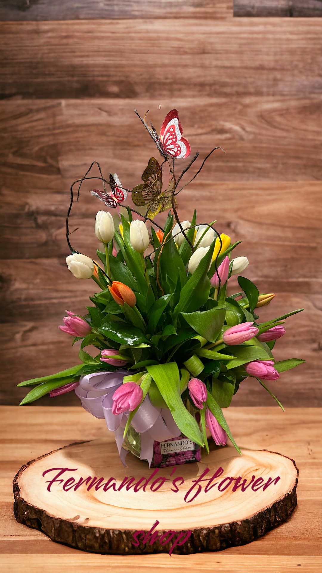 Mixed tulip arrangement with colorful butterfly picks in a clear vase on a wood slice base.