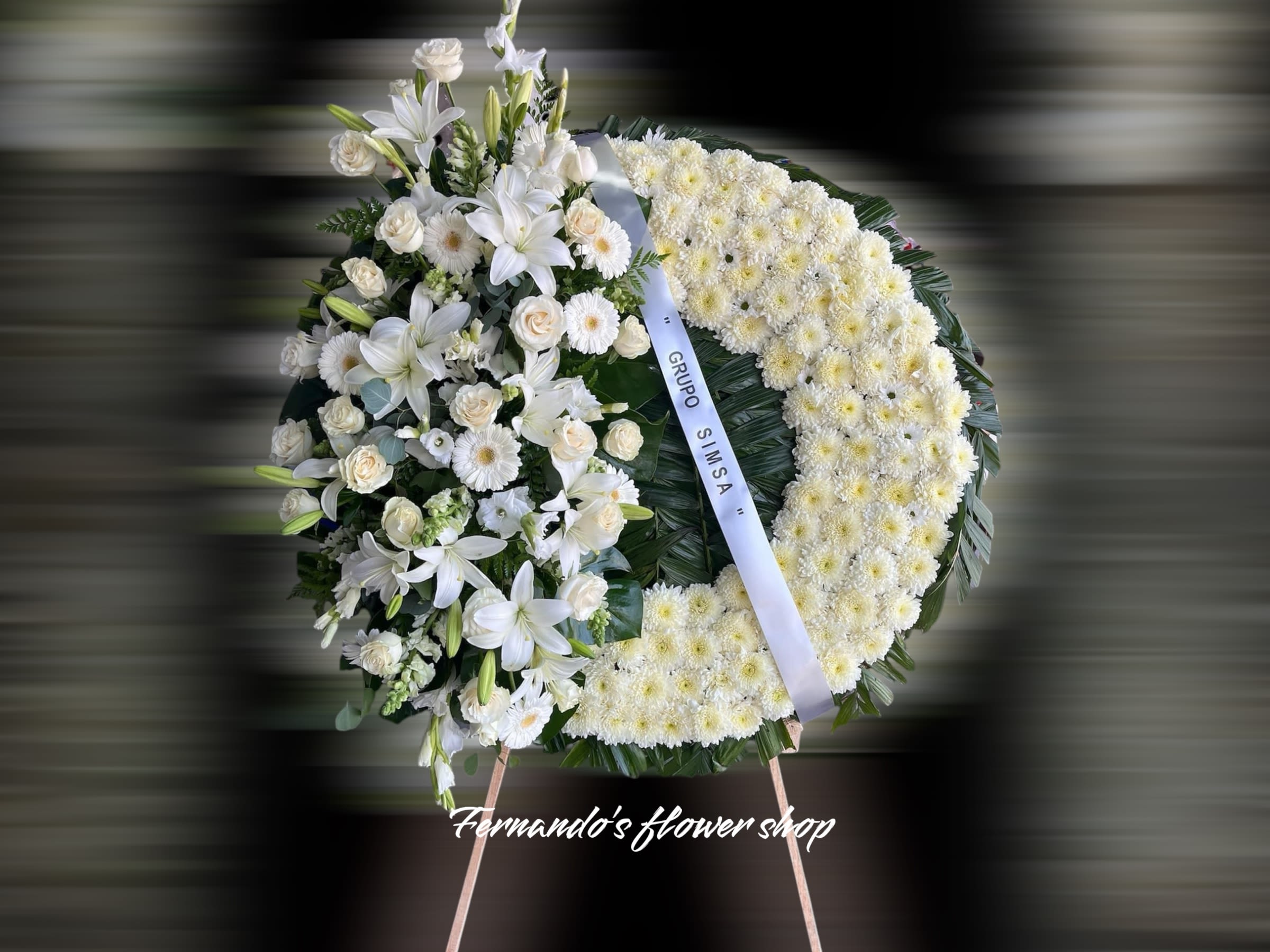 Standing white funeral wreath with lilies, roses, and chrysanthemums on an easel with a white ribbon