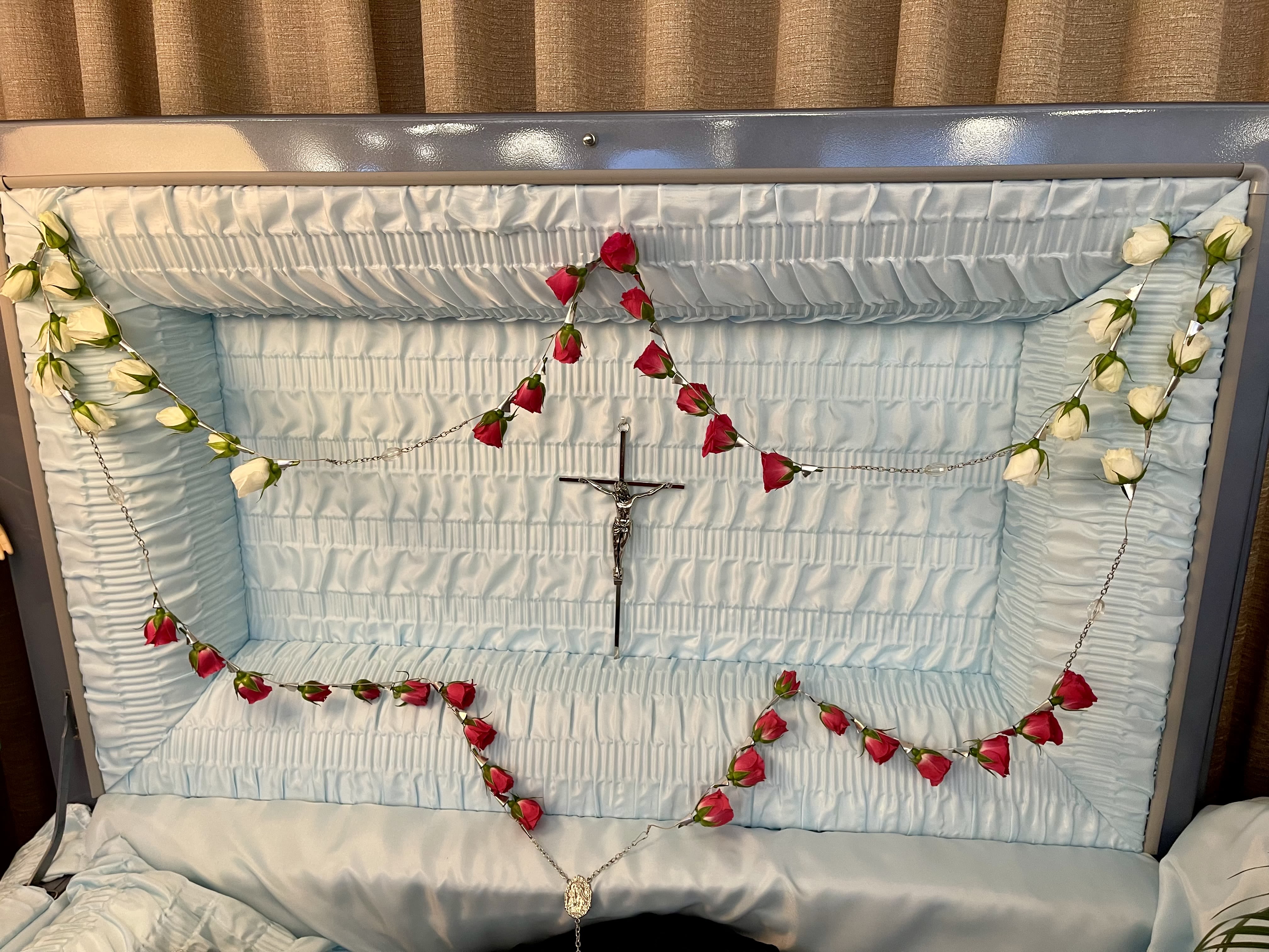 Red and white rosebud rosary draped inside a light blue open casket interior