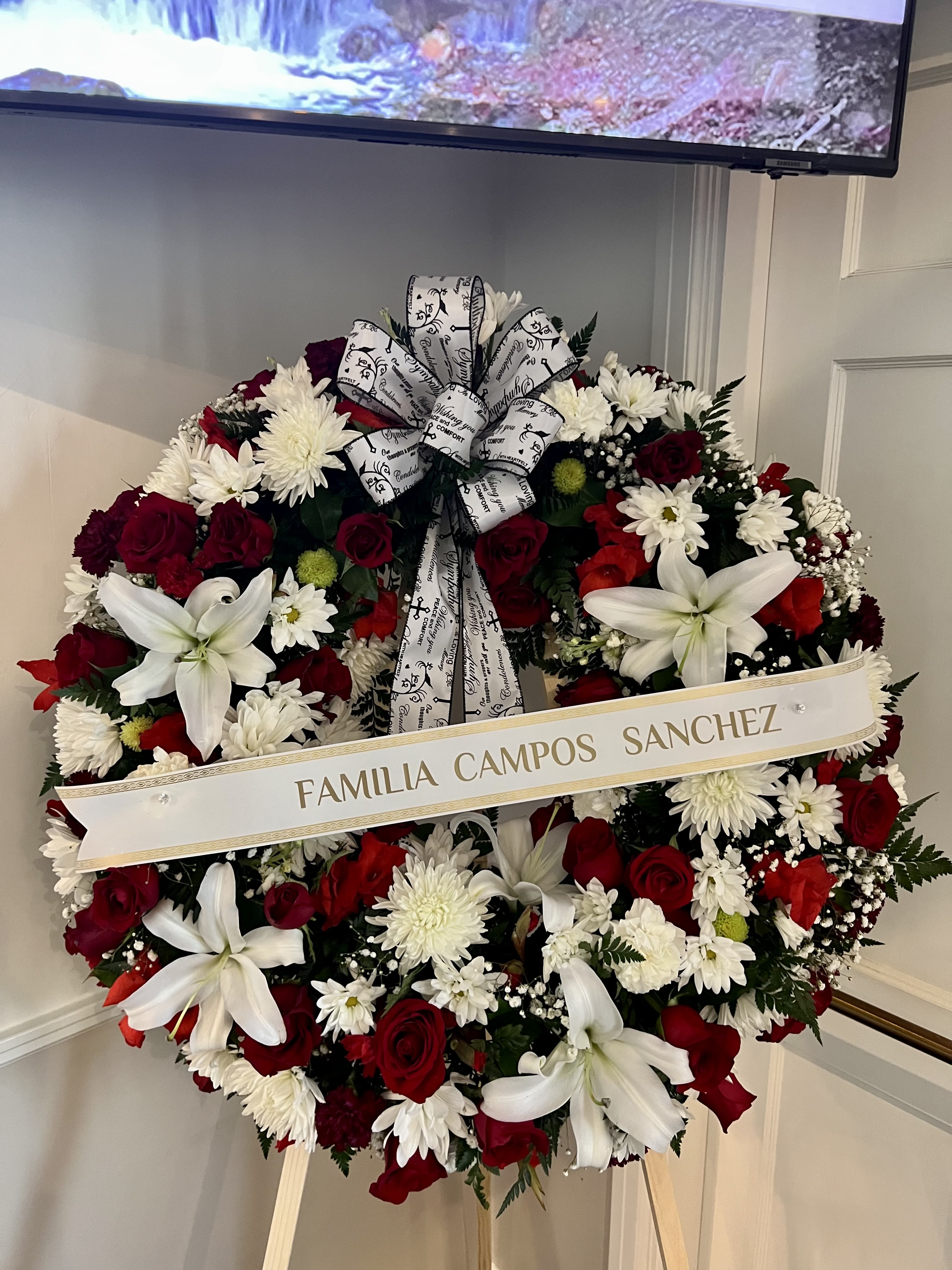 Standing round wreath of red roses, white lilies, and white mums with script ribbon on an easel
