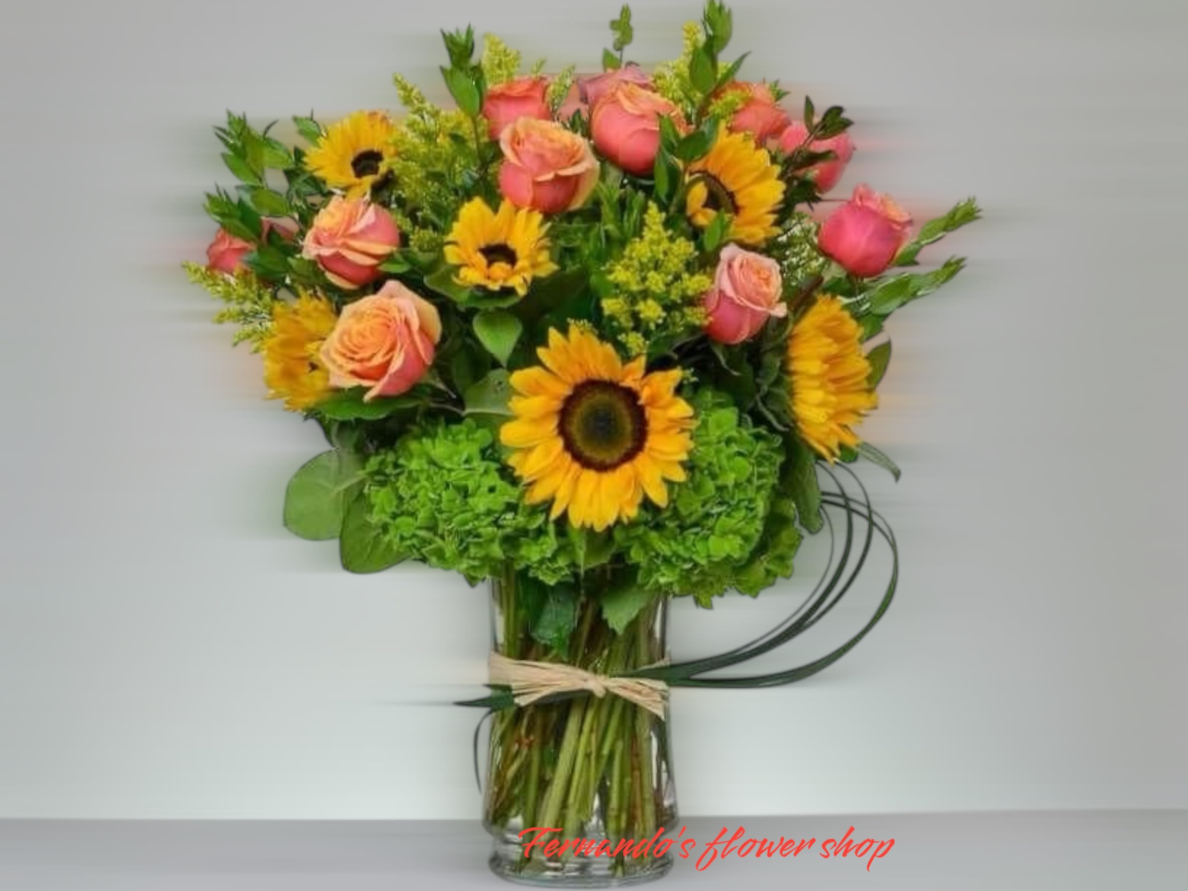 Vase arrangement of yellow sunflowers and pink roses with green hydrangeas in a clear glass cylinder