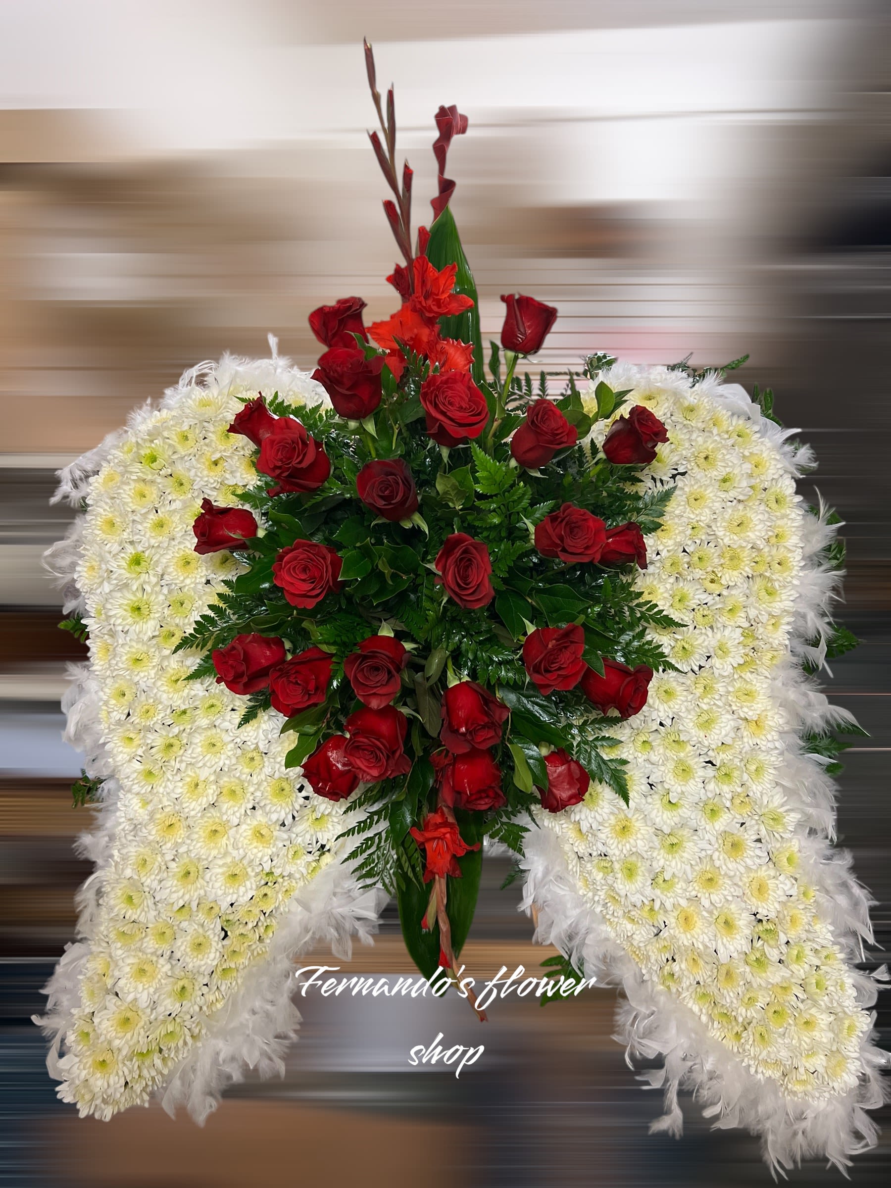 Fly High - This angelic floral arrangement features a pair of beautifully crafted wings made from delicate white chrysanthemums, symbolizing purity and peace. The wings are accented with a soft feather border, adding a heavenly and ethereal touch. At the center, vibrant red roses and gladiolus create a striking contrast, representing love and strength. Lush greenery enhances the overall design, providing depth and texture. This exquisite arrangement from “Fernando’s Flower Shop” is a perfect tribute for memorial services, offering a message of eternal love, hope, and remembrance.