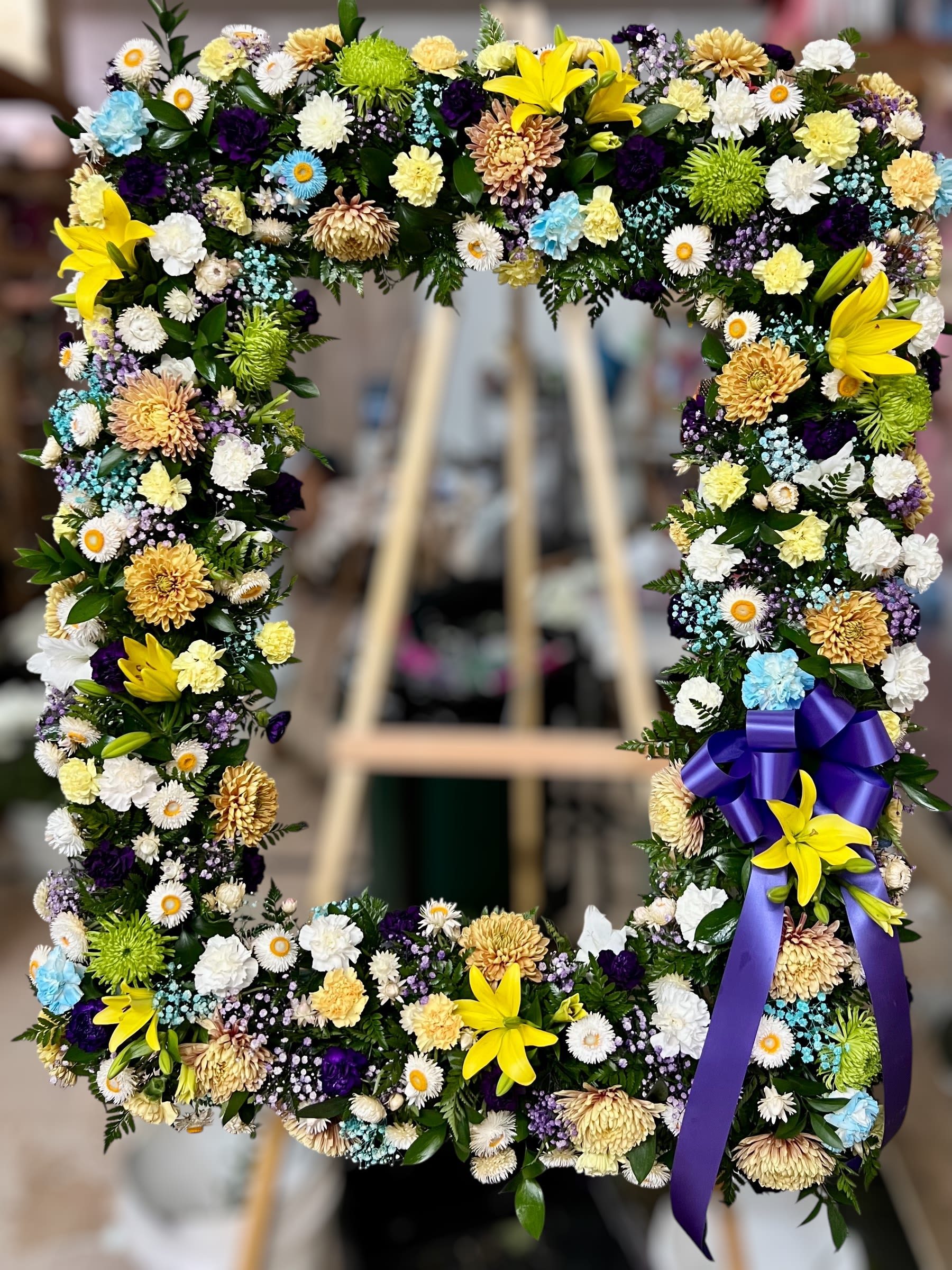 Rectangular standing wreath of yellow lilies, cream mums, and mixed small blooms with a purple bow