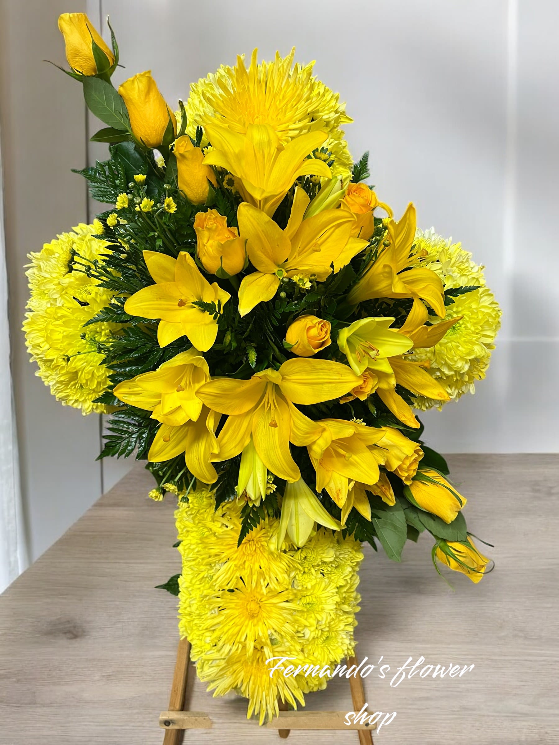 Yellow cross-shaped sympathy arrangement with lilies, roses, and chrysanthemums on a wooden stand