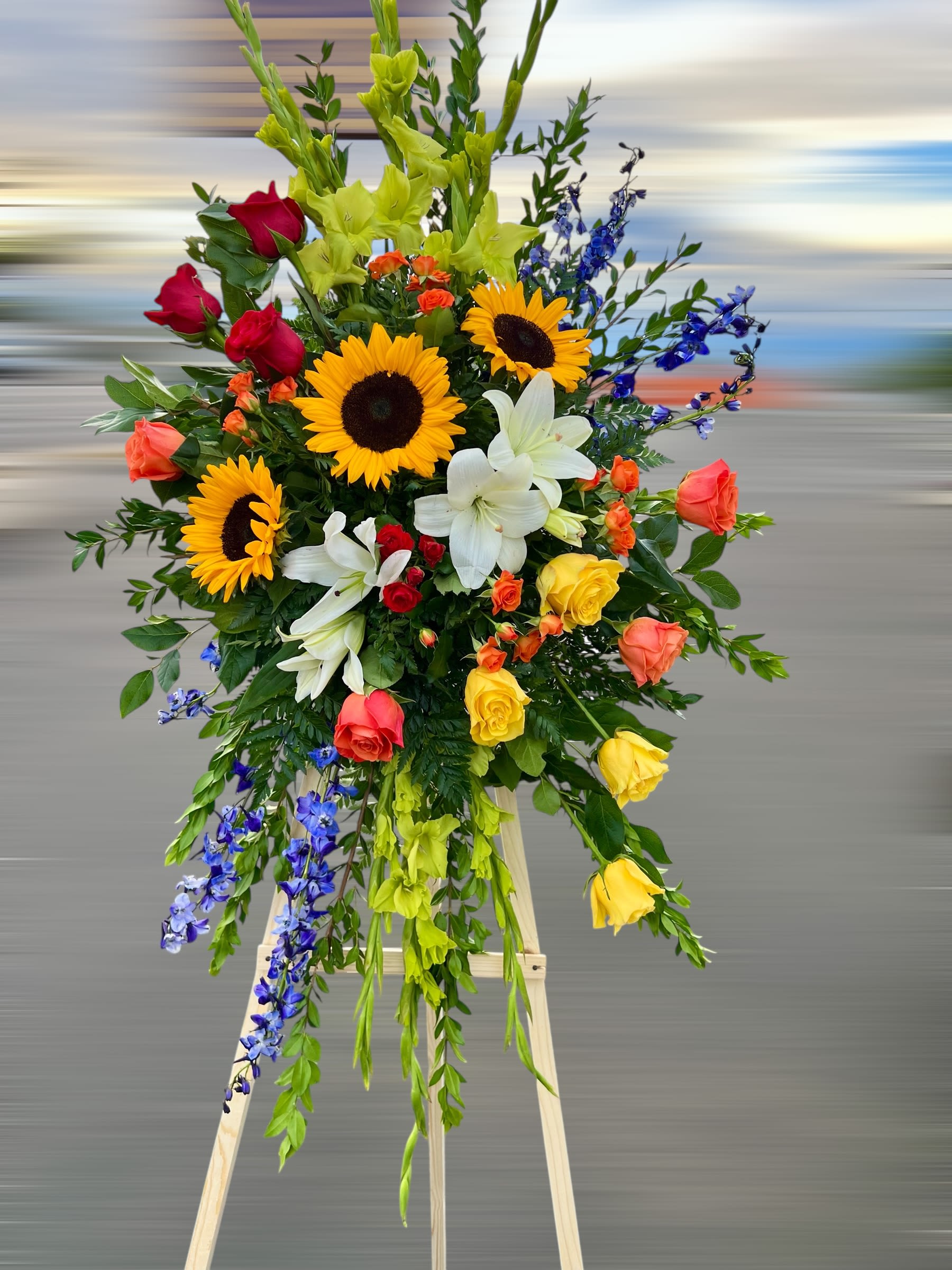 Tall standing spray of sunflowers, roses, lilies, and blue delphinium on a wooden easel