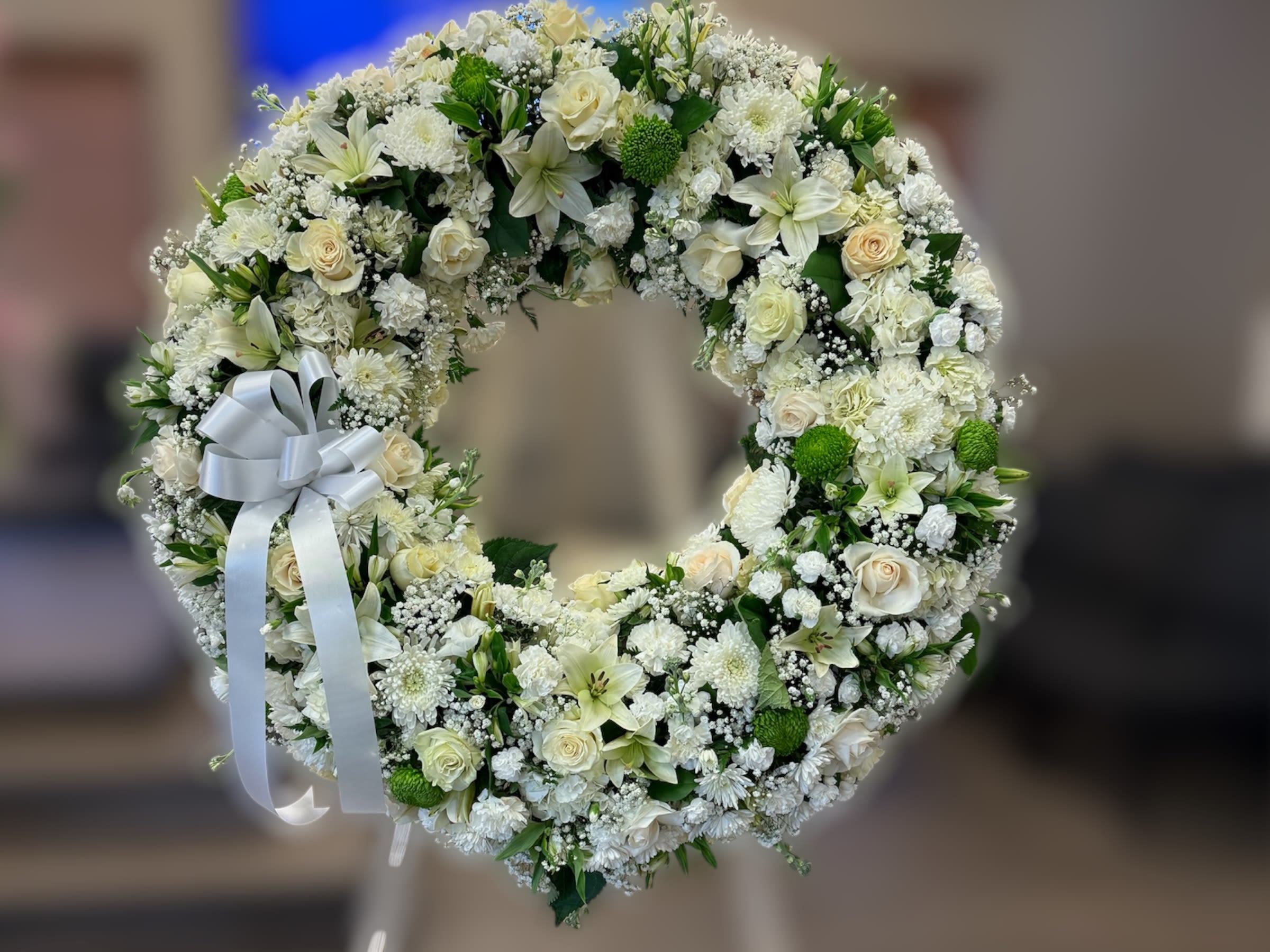 Large white and cream floral wreath with roses and lilies on a stand with a white bow