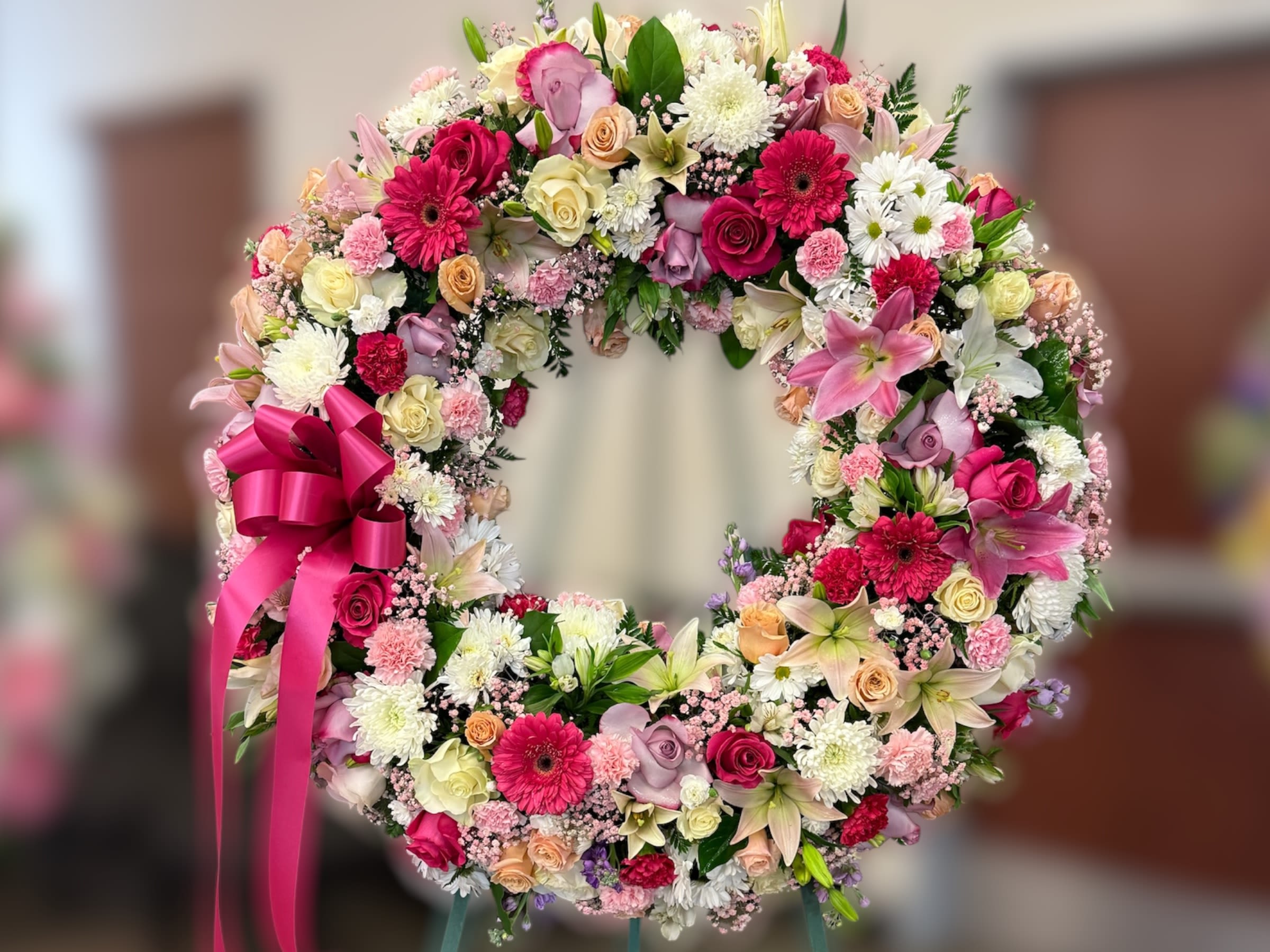 Standing wreath of pink, white, and peach flowers with a bright pink ribbon bow on an easel.
