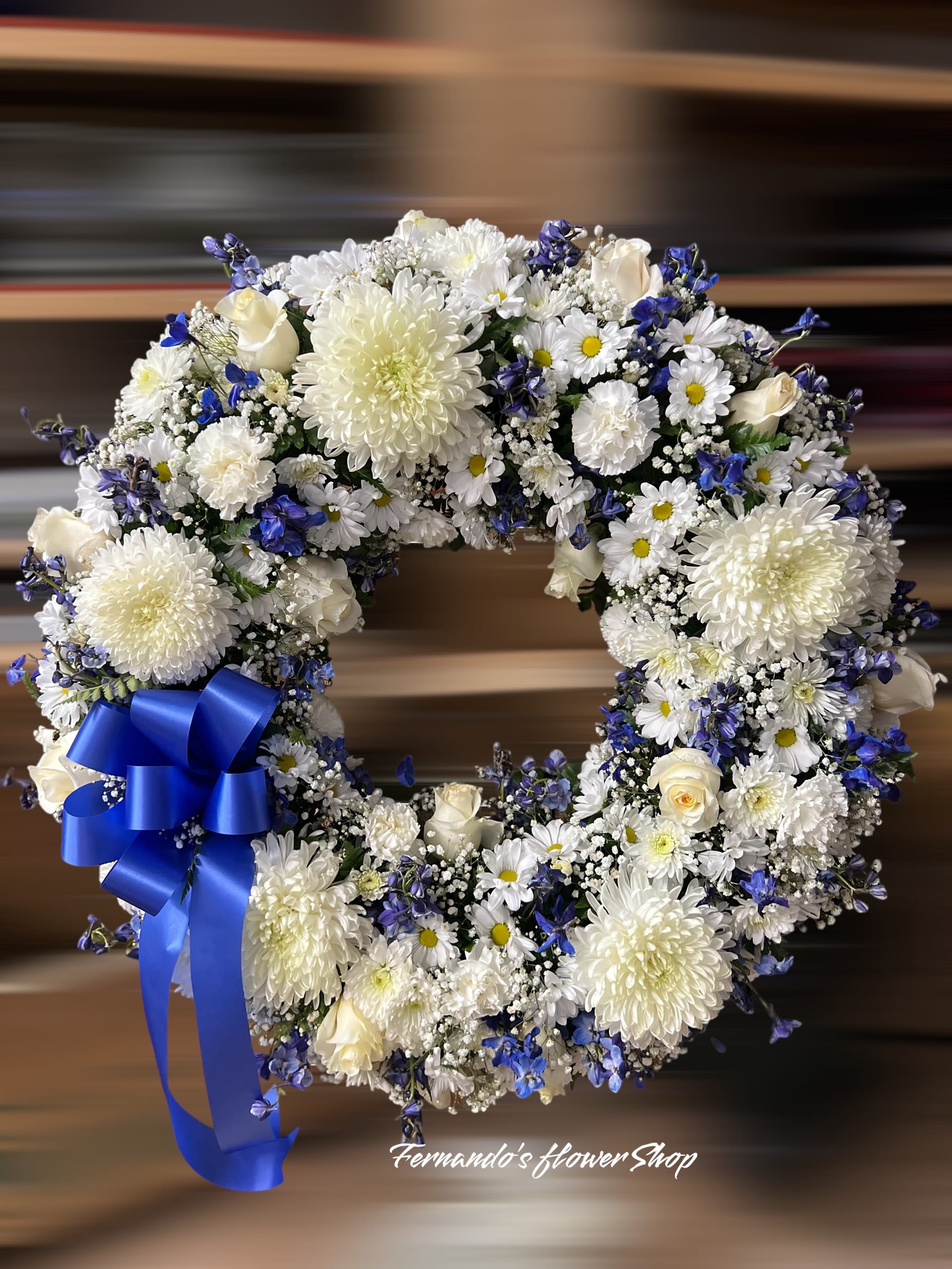 White and blue floral wreath with roses, daisies, and chrysanthemums, finished with a large royal blue bow.