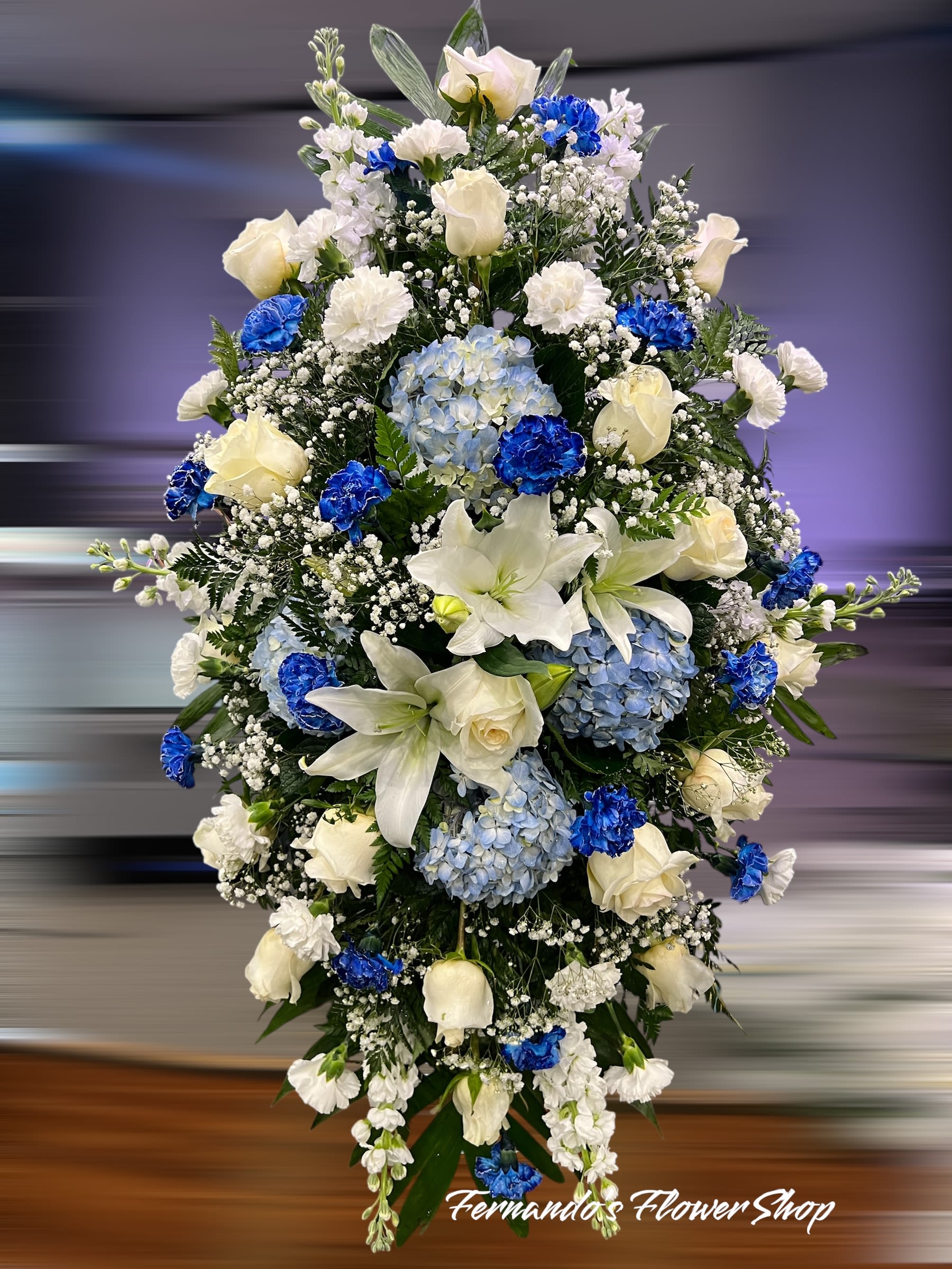 Tall standing spray of white lilies, roses, carnations and blue hydrangeas with bright blue accent blooms.
