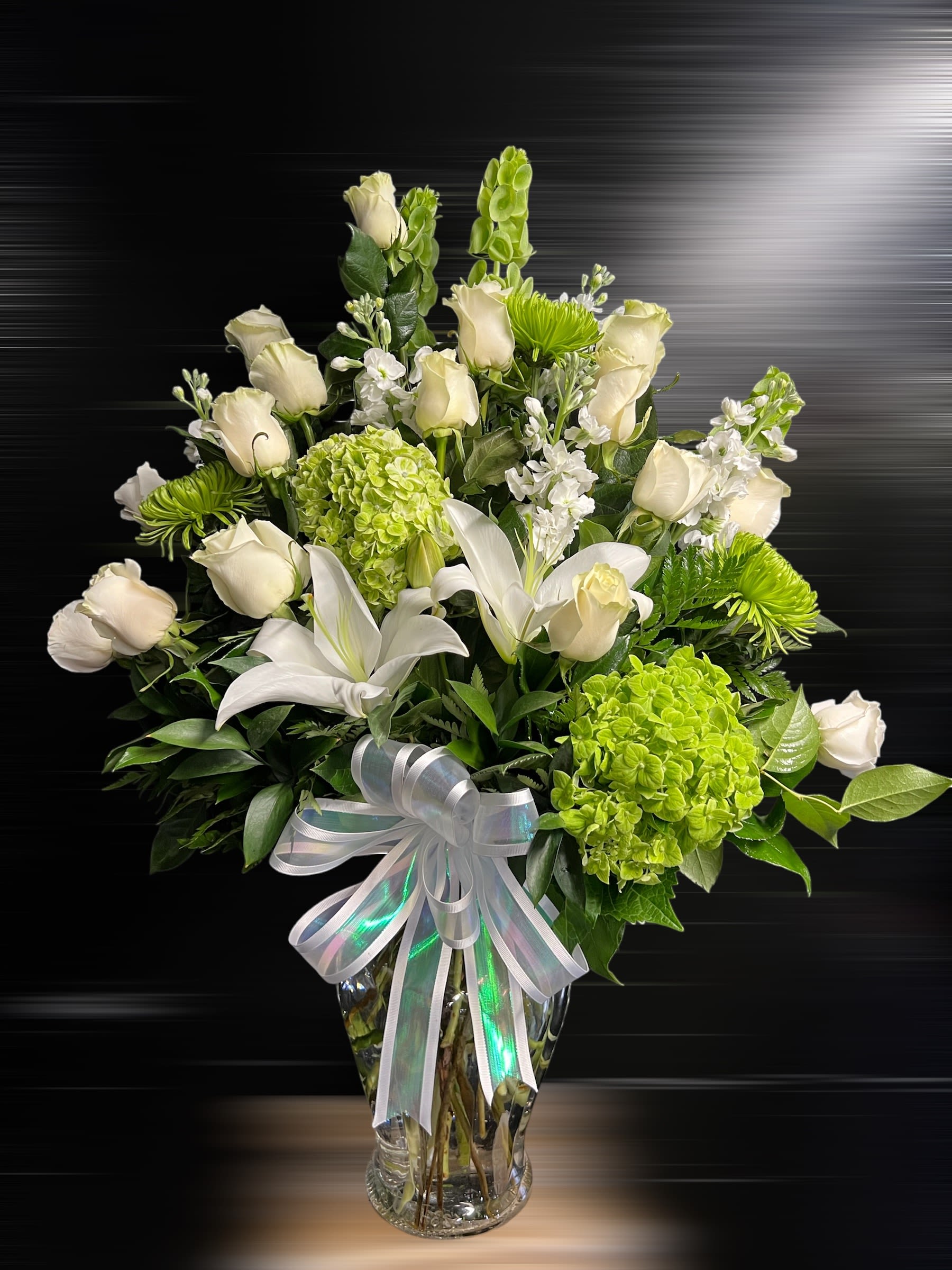 Tall vase arrangement of white roses and lilies with green hydrangeas and mums, finished with a white bow.