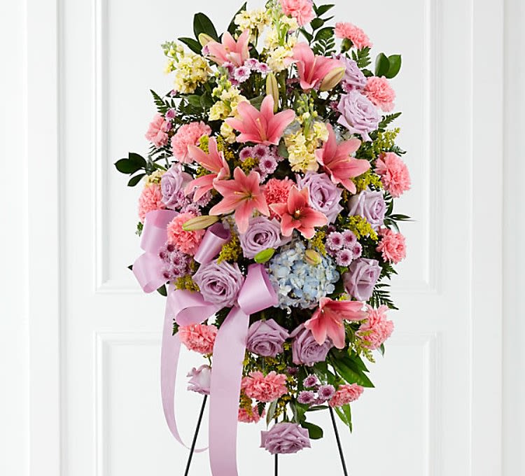 Standing spray of pink lilies, lavender roses, and carnations on an easel with lavender ribbon.