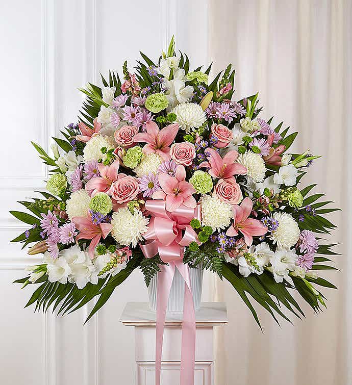 Fan-shaped pastel arrangement with pink lilies and roses, white mums, and a pink ribbon in a white container
