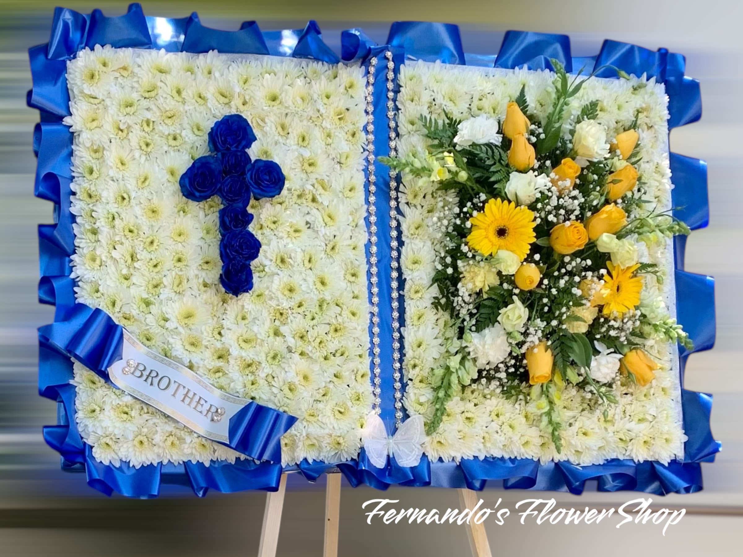 Standing open-book funeral arrangement with blue rose cross and yellow and white flowers.