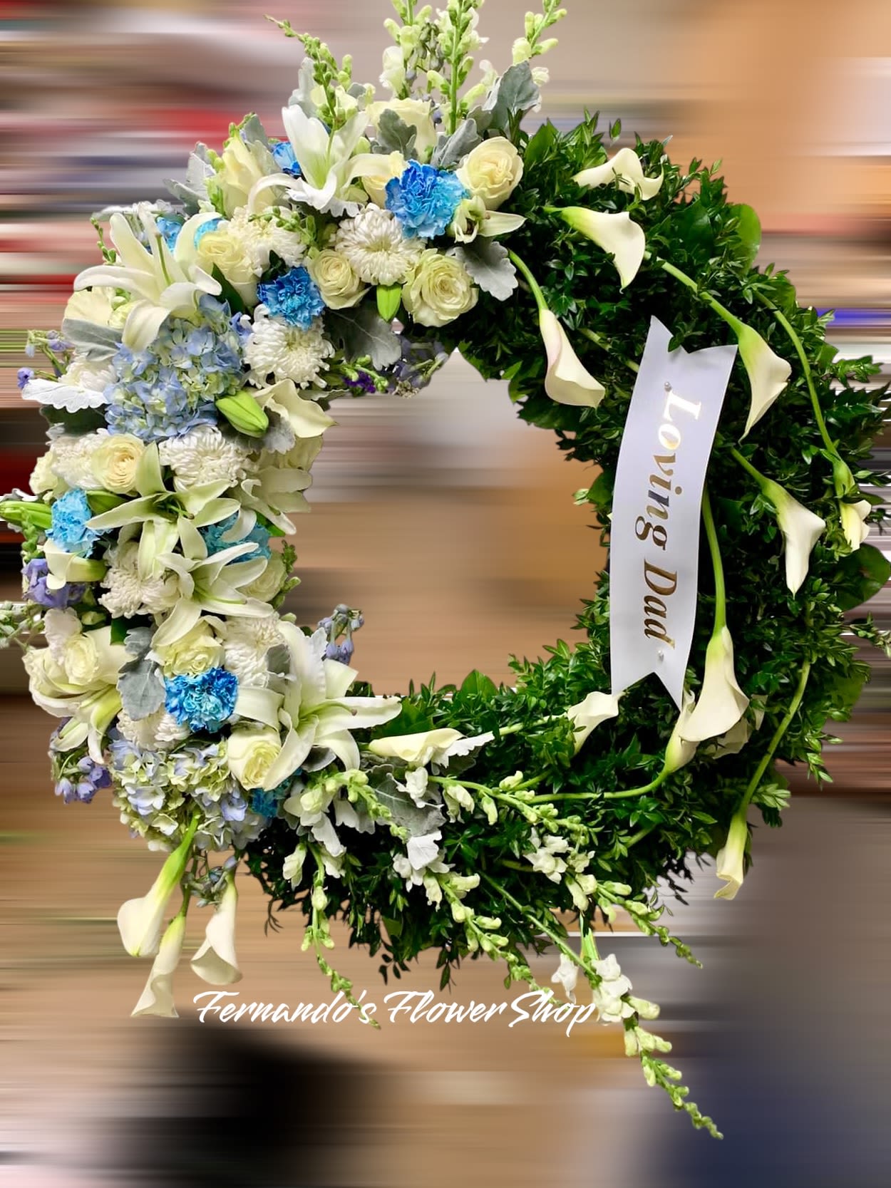 Round sympathy wreath with white lilies, roses, blue flowers, and a Loving Dad ribbon.