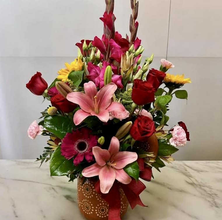 Tall mixed arrangement with pink lilies, red roses, yellow blooms in an orange patterned vase with ribbon
