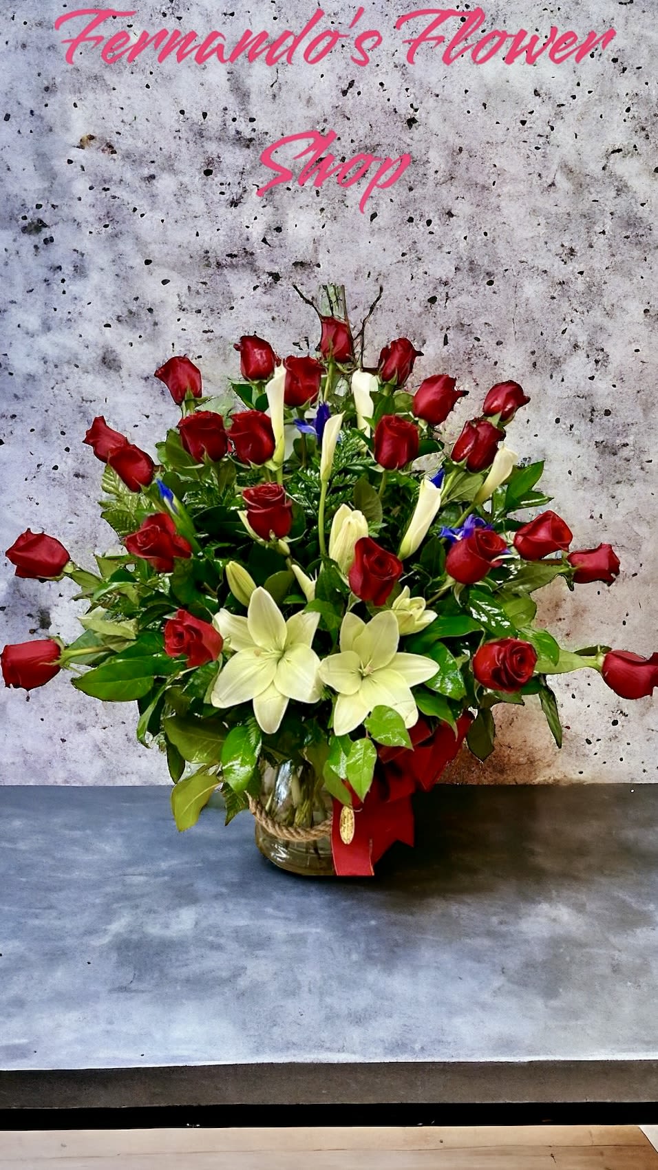 Large arrangement of red roses and white lilies with greenery in a clear glass vase tied with rope and a red bow
