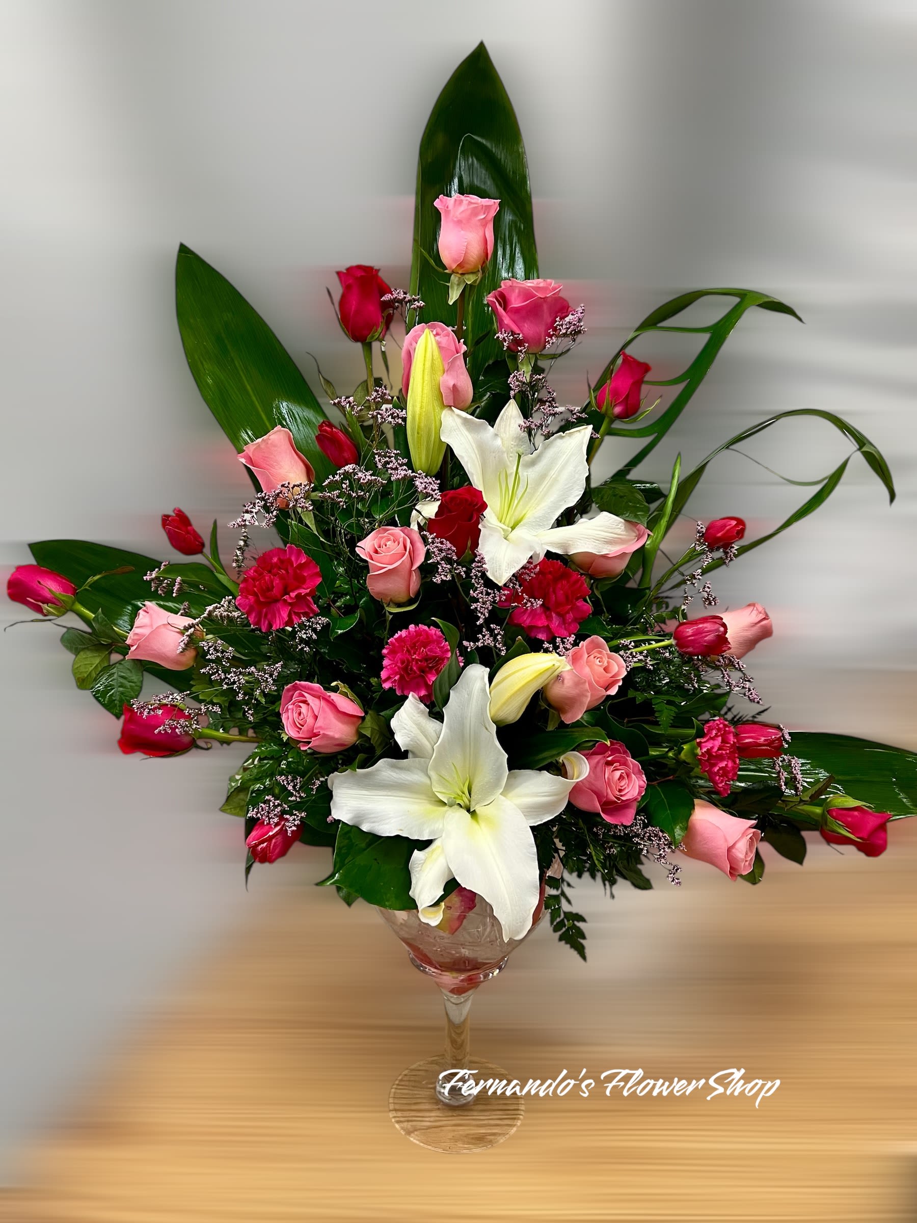 Tall arrangement of pink and red roses, white lilies, and bright pink carnations in a clear glass goblet vase