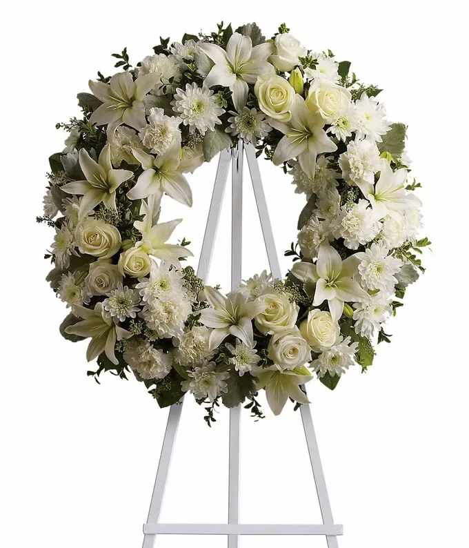 White floral wreath of lilies, roses, and mums displayed on a white easel stand