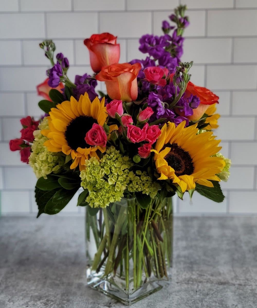 Bright mixed arrangement with sunflowers, roses, and purple blooms in a clear glass vase