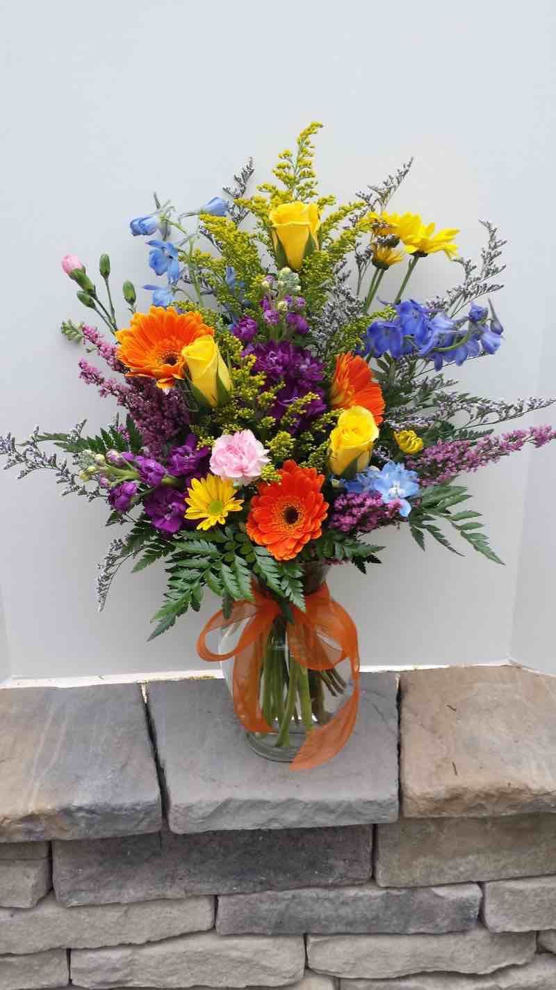 Tall mixed arrangement of bright yellow roses, orange gerberas, and purple and blue flowers in a glass vase