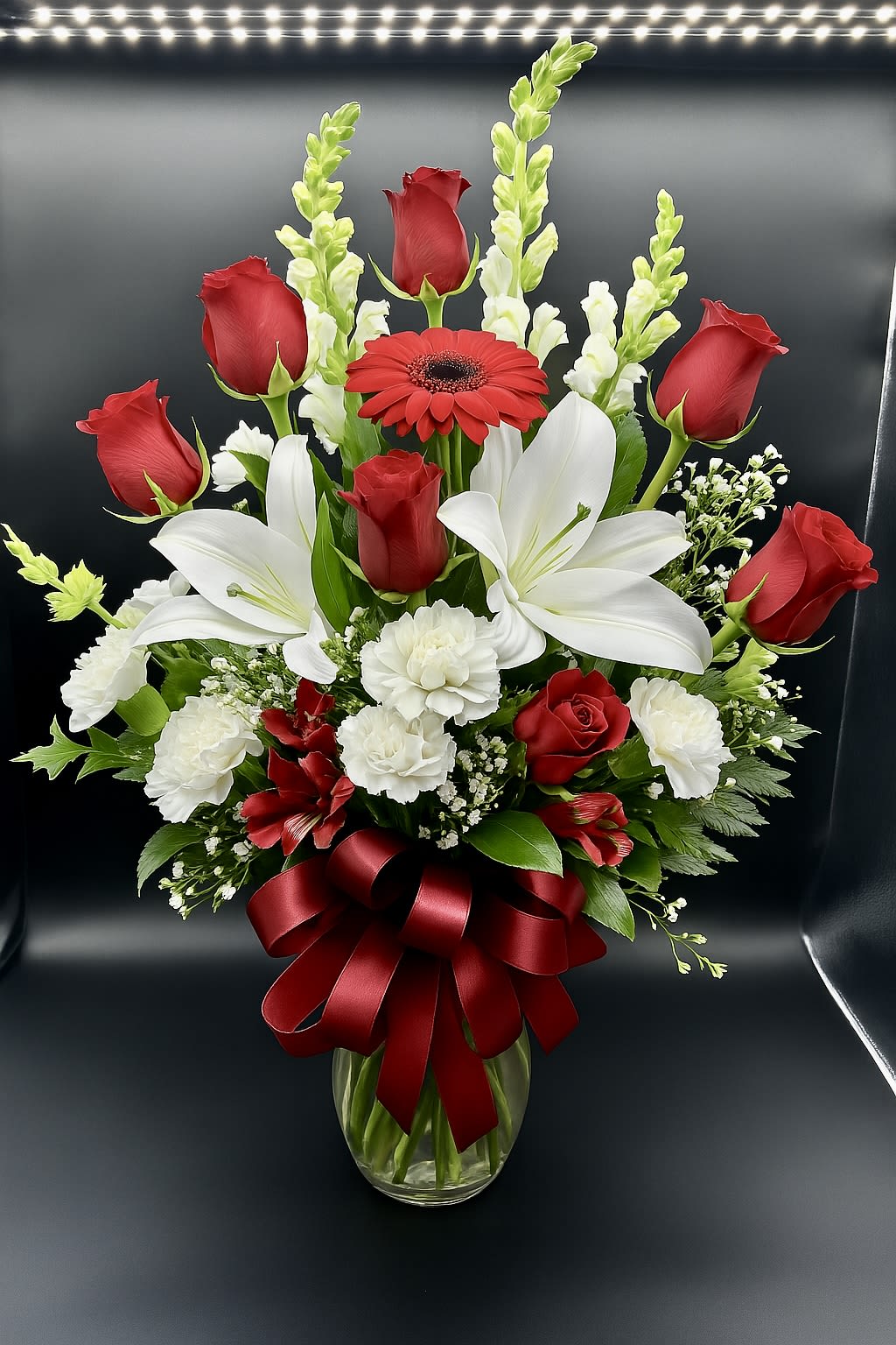 Tall arrangement of red roses, white lilies, and carnations in a glass vase with a red bow
