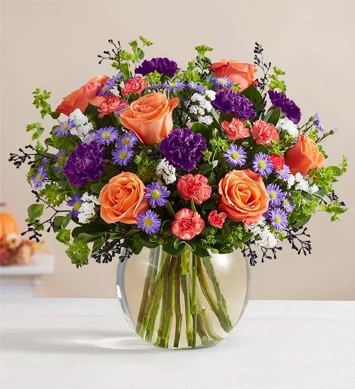 Mixed bouquet of orange roses, purple carnations, and small daisies in a round clear glass vase