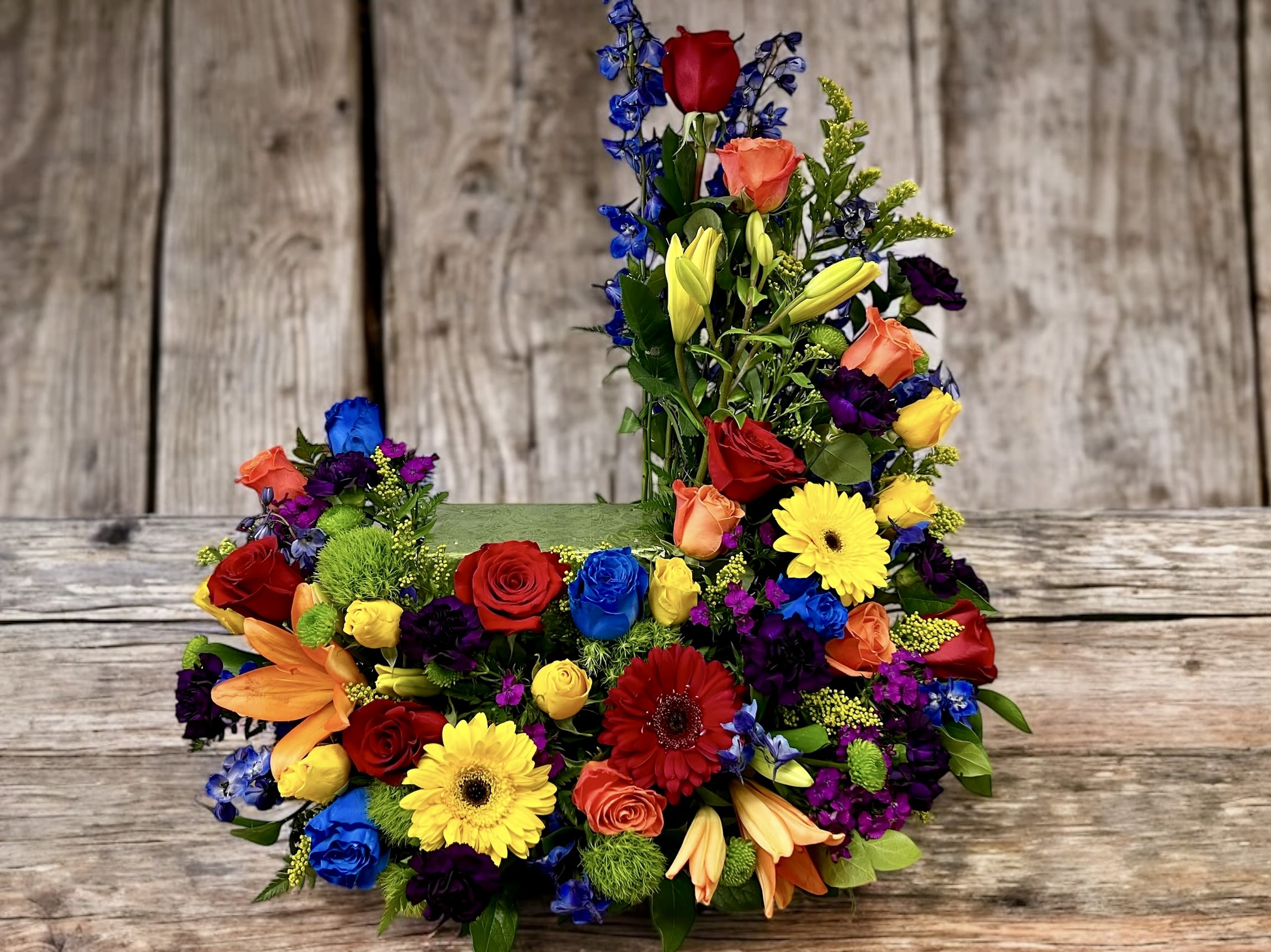 Tall and low arrangement of bright roses, lilies, gerbera daisies, and other blooms in a rectangular container.