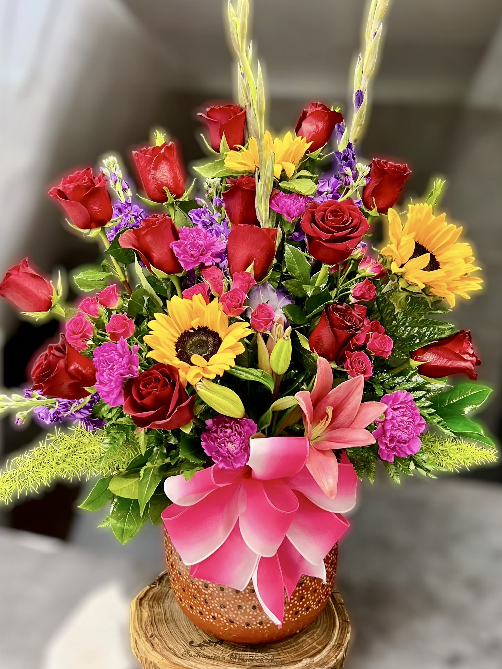Tall mixed arrangement with red roses, sunflowers, pink lilies, and carnations in an orange pot with a pink bow.