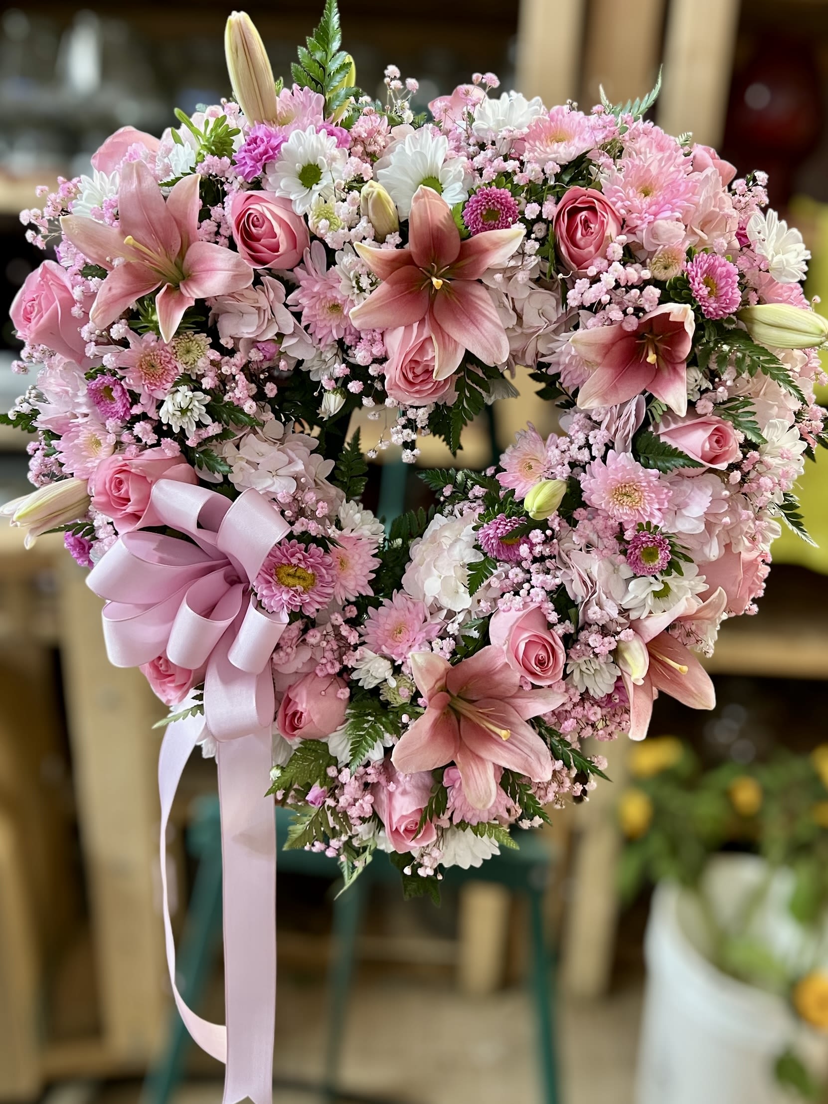 Heart-shaped standing wreath of pink lilies, roses, and mums with a large pink ribbon bow.