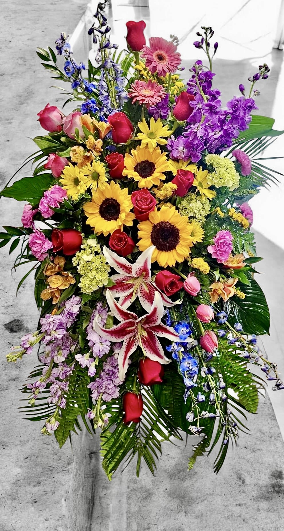 Tall standing spray of mixed bright flowers including sunflowers, roses, lilies, and gerbera daisies with palm fronds