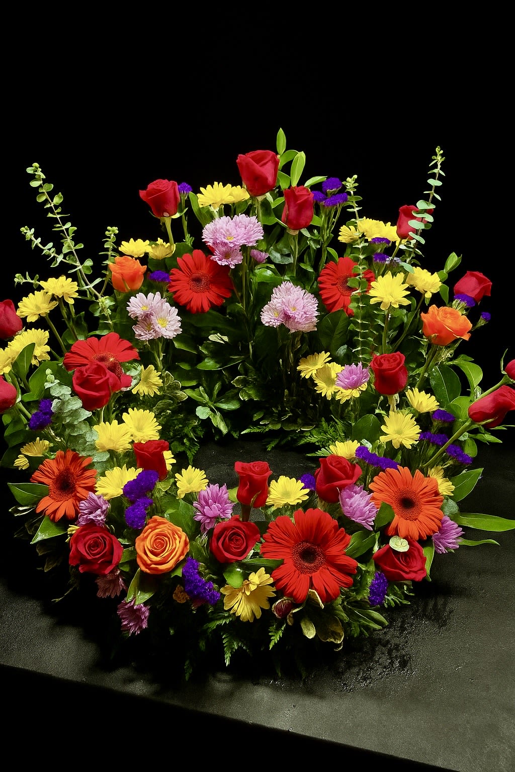 Bright mixed flower wreath of roses, gerbera daisies, and mums in a circular design on a dark background
