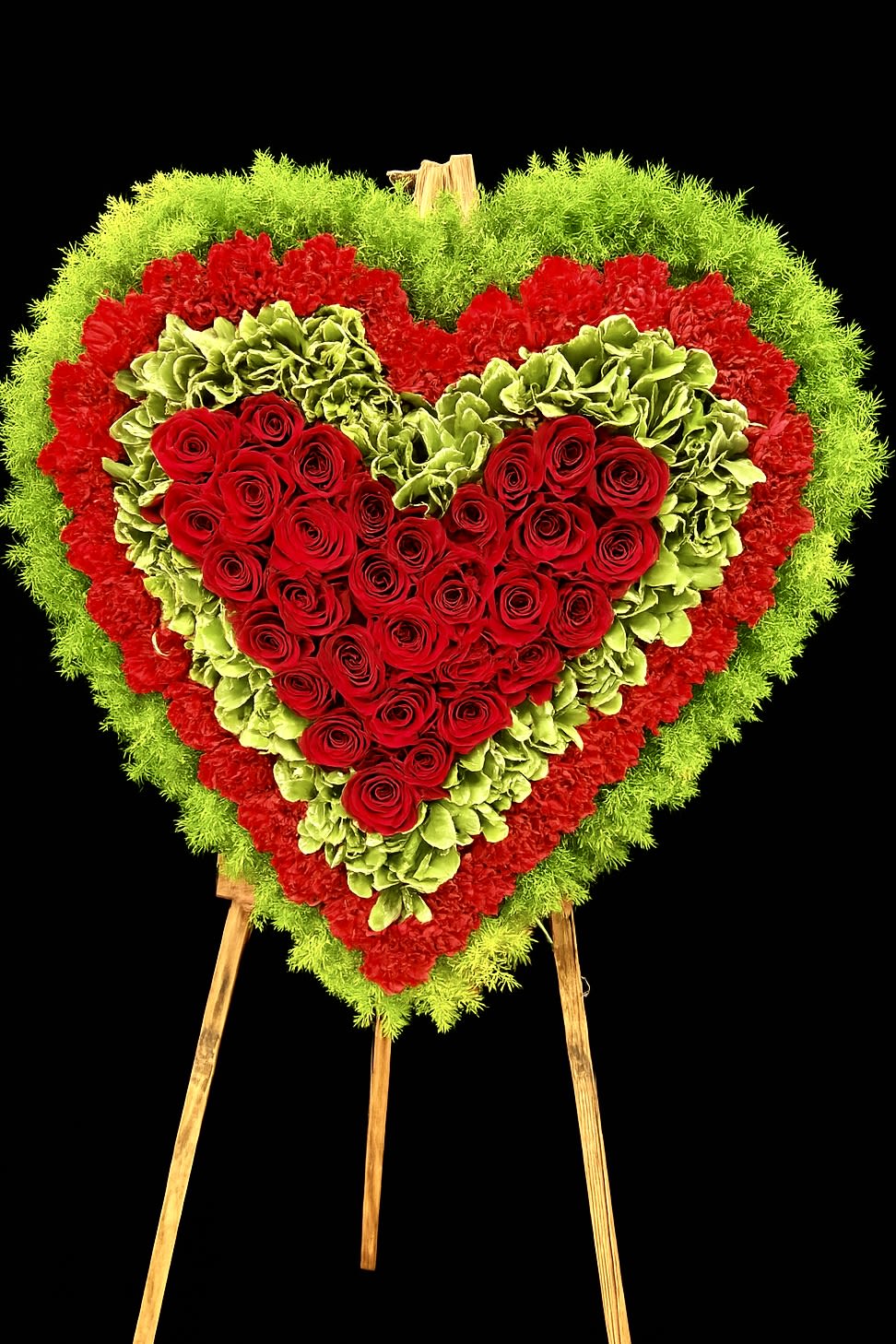 Heart-shaped standing floral spray of red roses with green and red flower borders on a wooden easel.