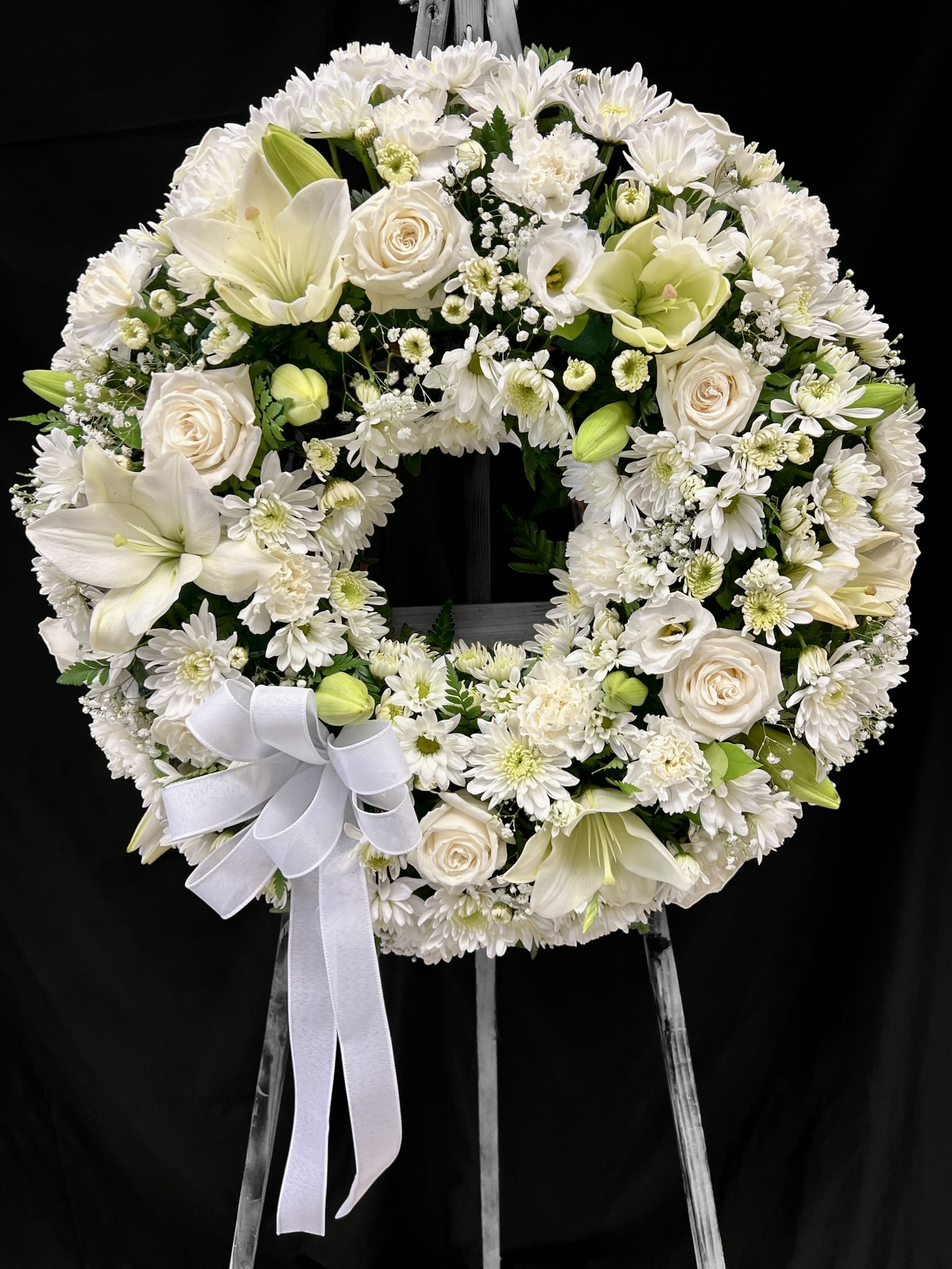 Standing white funeral wreath of roses, lilies, and chrysanthemums with a bow on a metal easel