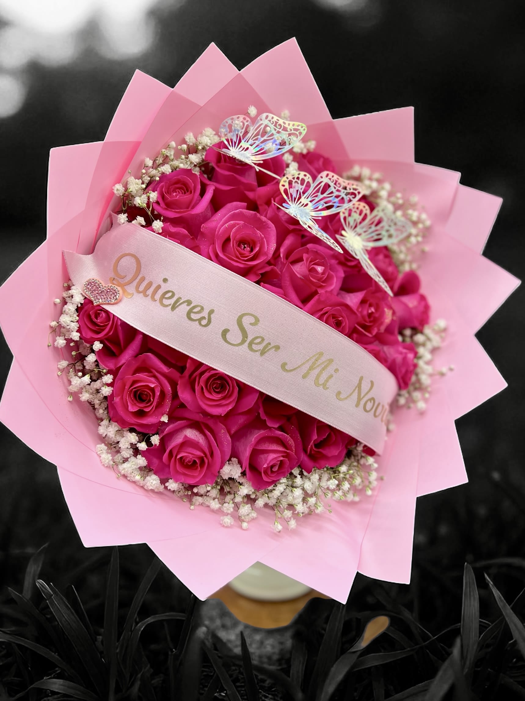 Bouquet of bright pink roses with baby's breath, pink wrapping, and a ribbon with Spanish proposal text