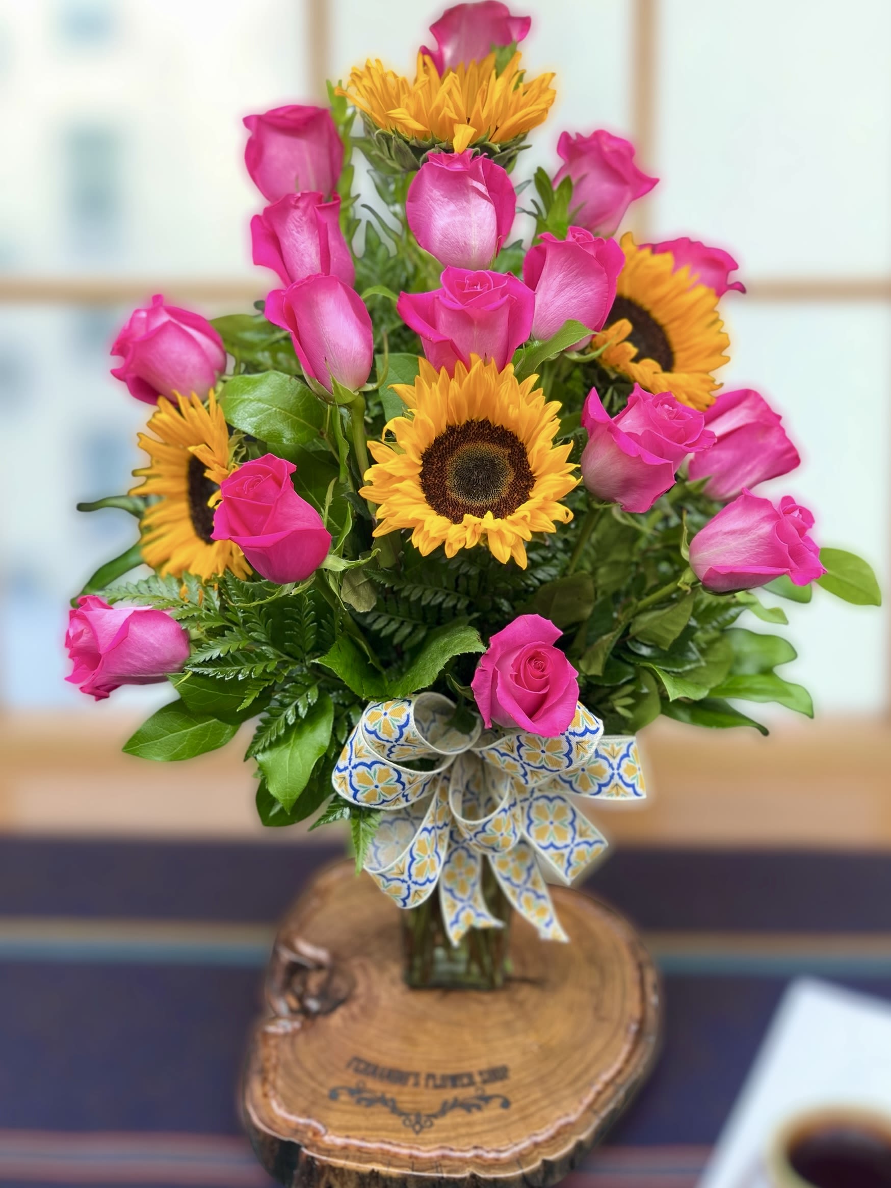 Arrangement of bright pink roses and yellow sunflowers in a glass vase with a patterned ribbon bow
